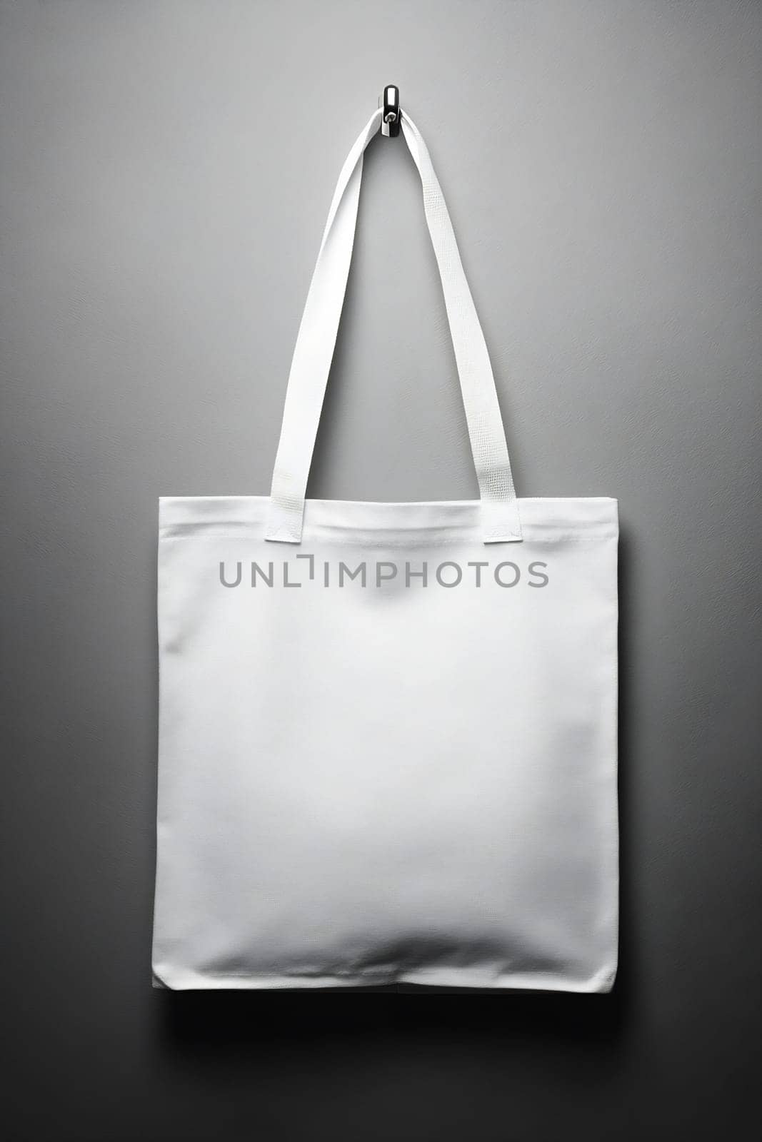 A plain white tote bag is suspended from a hook against a neutral background, showcasing its minimalist design and versatility for use.