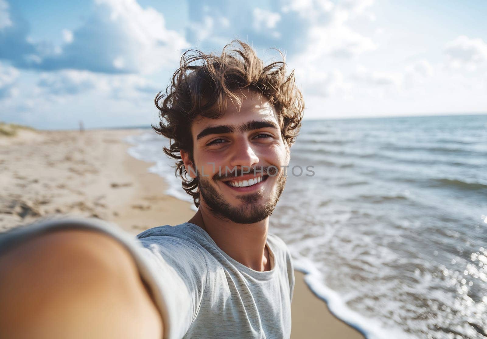 A young man smiles at the camera while standing on a beach, with waves gently lapping at the shore during a sunny day.