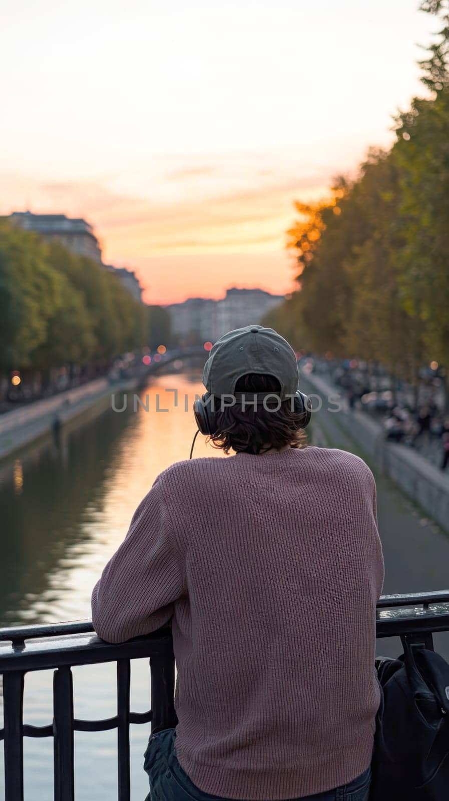 A young man leaning against a wall near the canals, wearing an oversized sweater and a baseball cap, listening to music through his vintage headphones. AI generative.