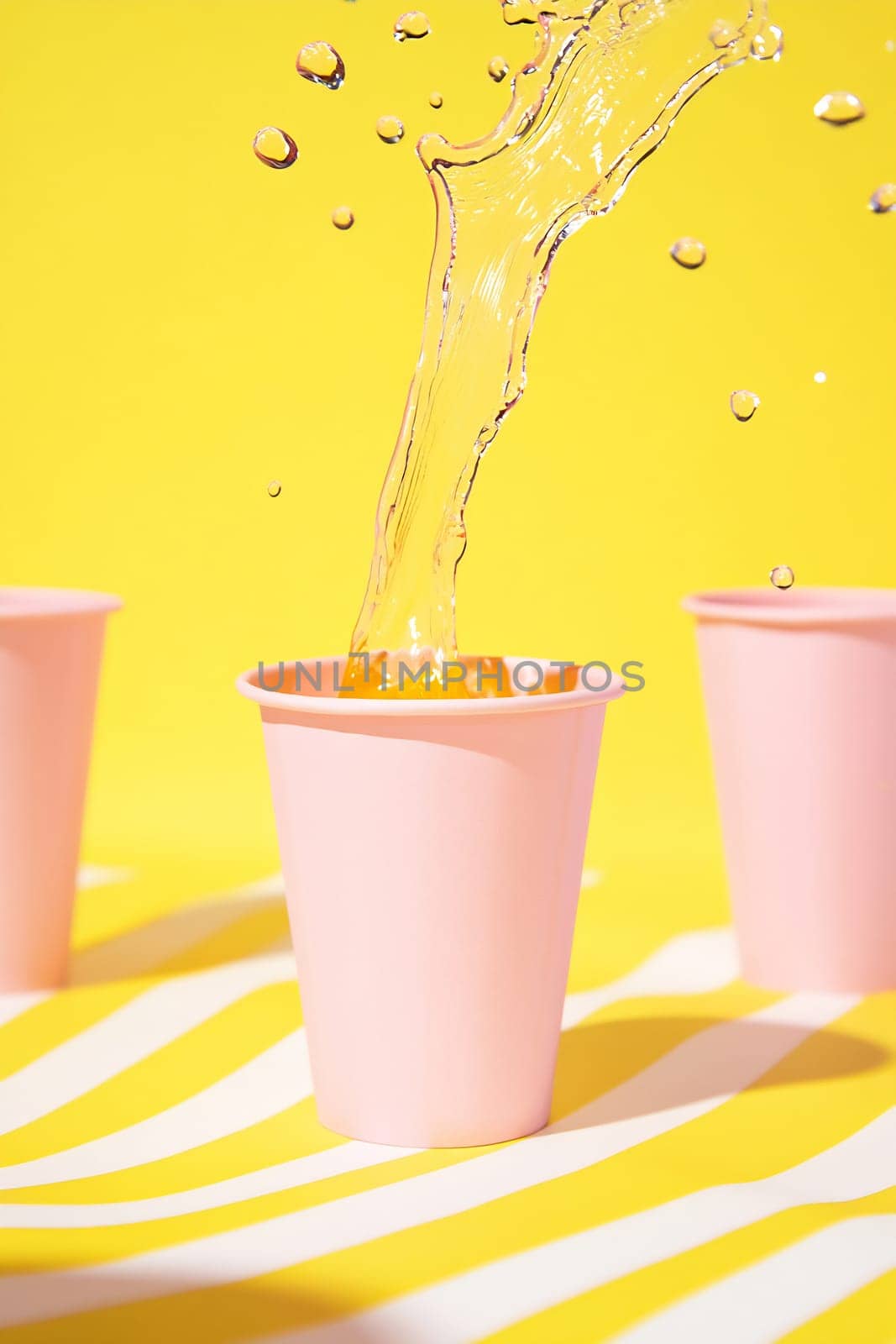 A colorful drink is splashed into a pink cup, surrounded by more cups against a bright yellow background.