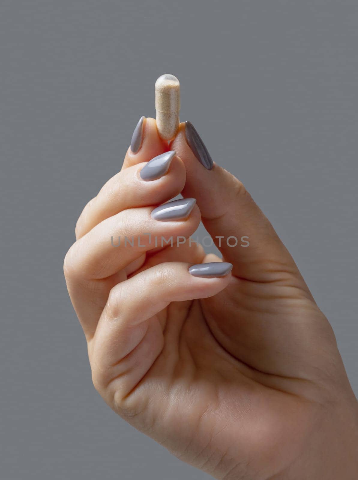 Female hand holding a beige capsule with fingers closeup against grey background. Healthcare, medical and pharmaceutical concept, herbal alternative cure