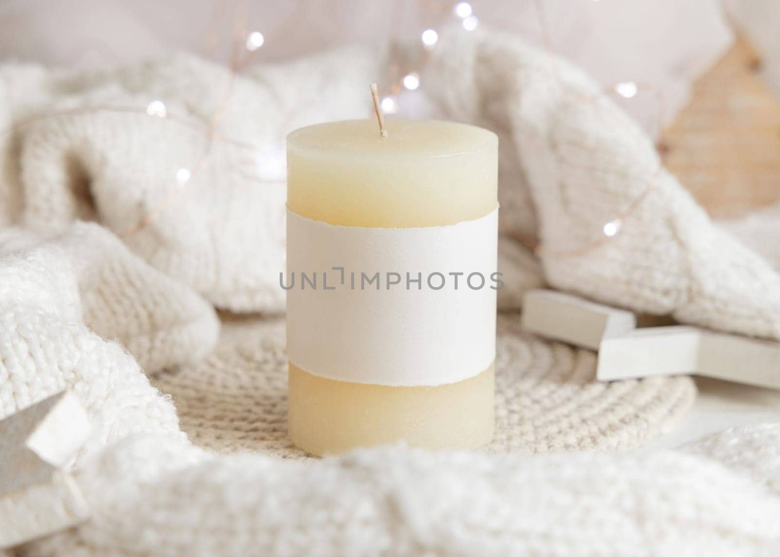 Pillar candle with blank label near white knitted sweater, wooden Christmas decor and lights, copy space. Winter atmospheric mock up in rustic or scandinavian style