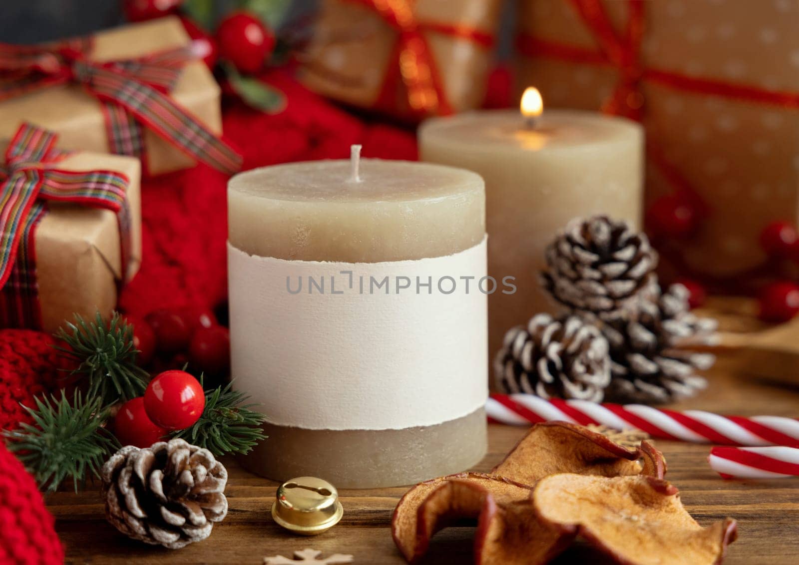 Pillar candles with blank label near red knitted sweater and Christmas decor against lights and wrapped presents, copy space. Warm winter family atmospheric mock up, Holiday time