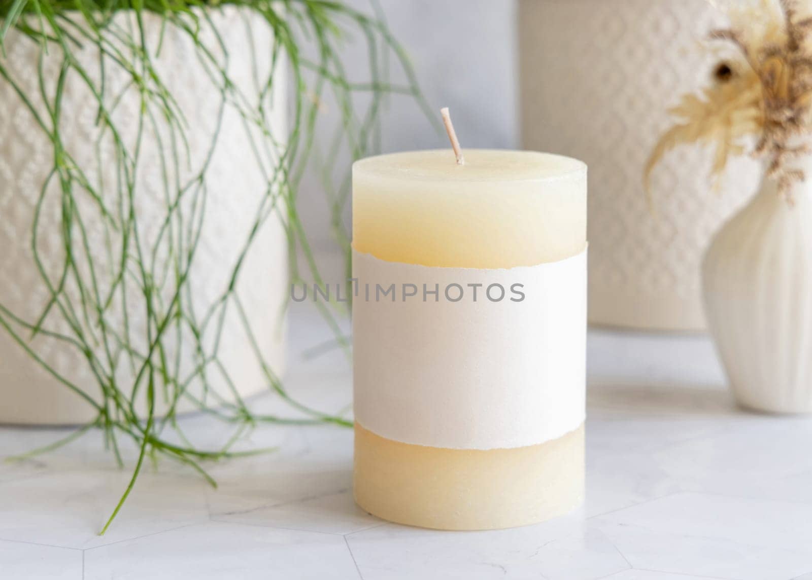 Pillar candle with blank label on marble near potted green plant close up, packaging mockup. Minimal scandinavian home decoration, romantic interior