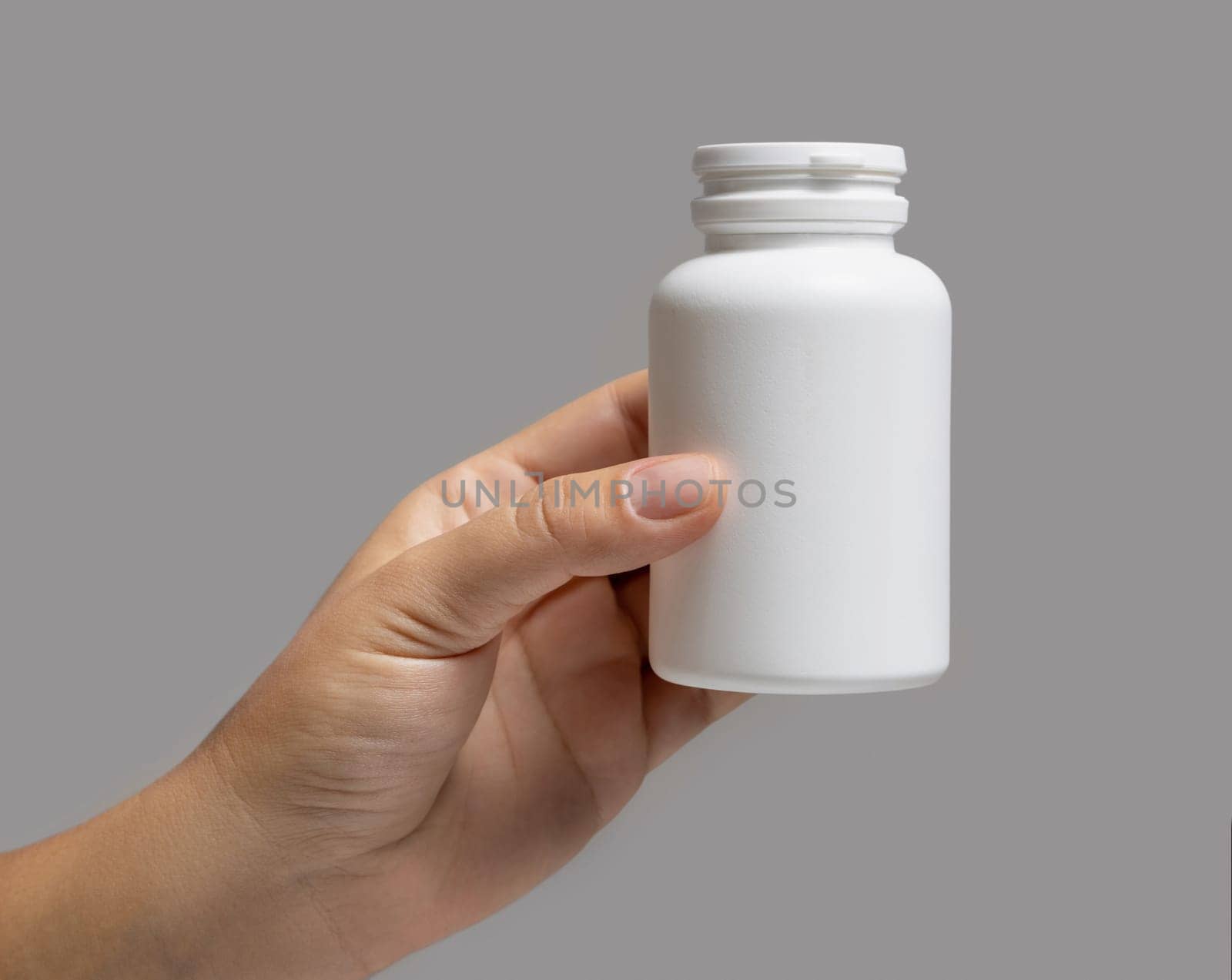 Female hand holding a white plastic pill bottle closeup against grey background. Packaging mockup for your pills or capsules herbal supplement