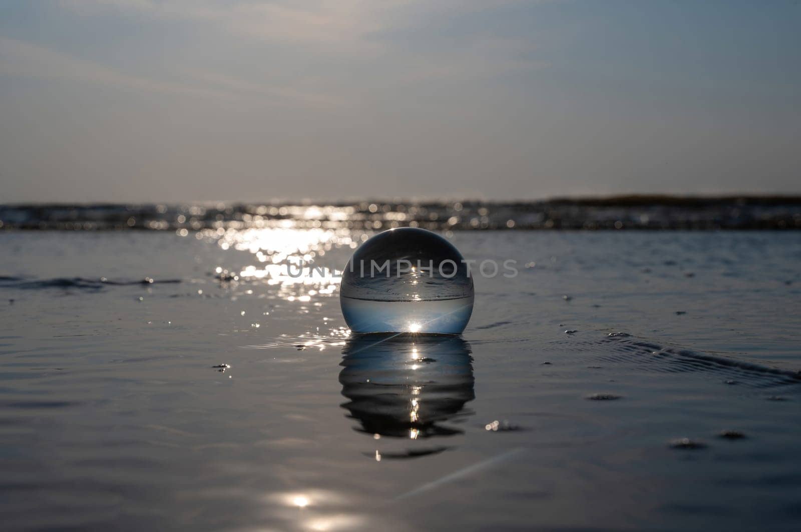A glass ball lies in the waves on the sandy beach, the sea and the setting sun are reflected in the ball
