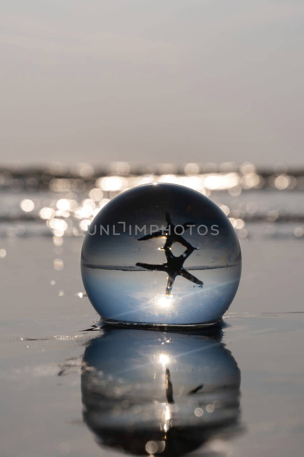 A glass ball lies in the waves on the sandy beach, the ball reflects the sea, a starfish and the setting sun