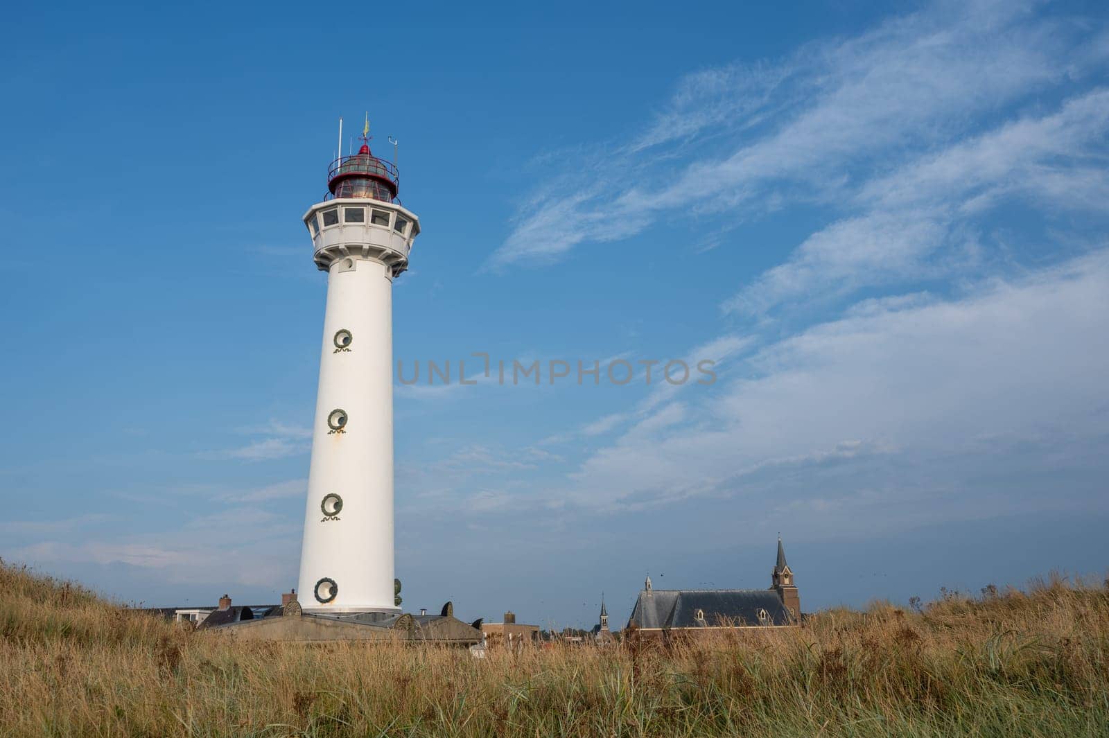 The approx. 28 m high white lighthouse J.C.J. van Speijk in Egmond aan Zee, North Holland, Netherlands with the Catholic St. Agnes Church, beach grass and sand dunes