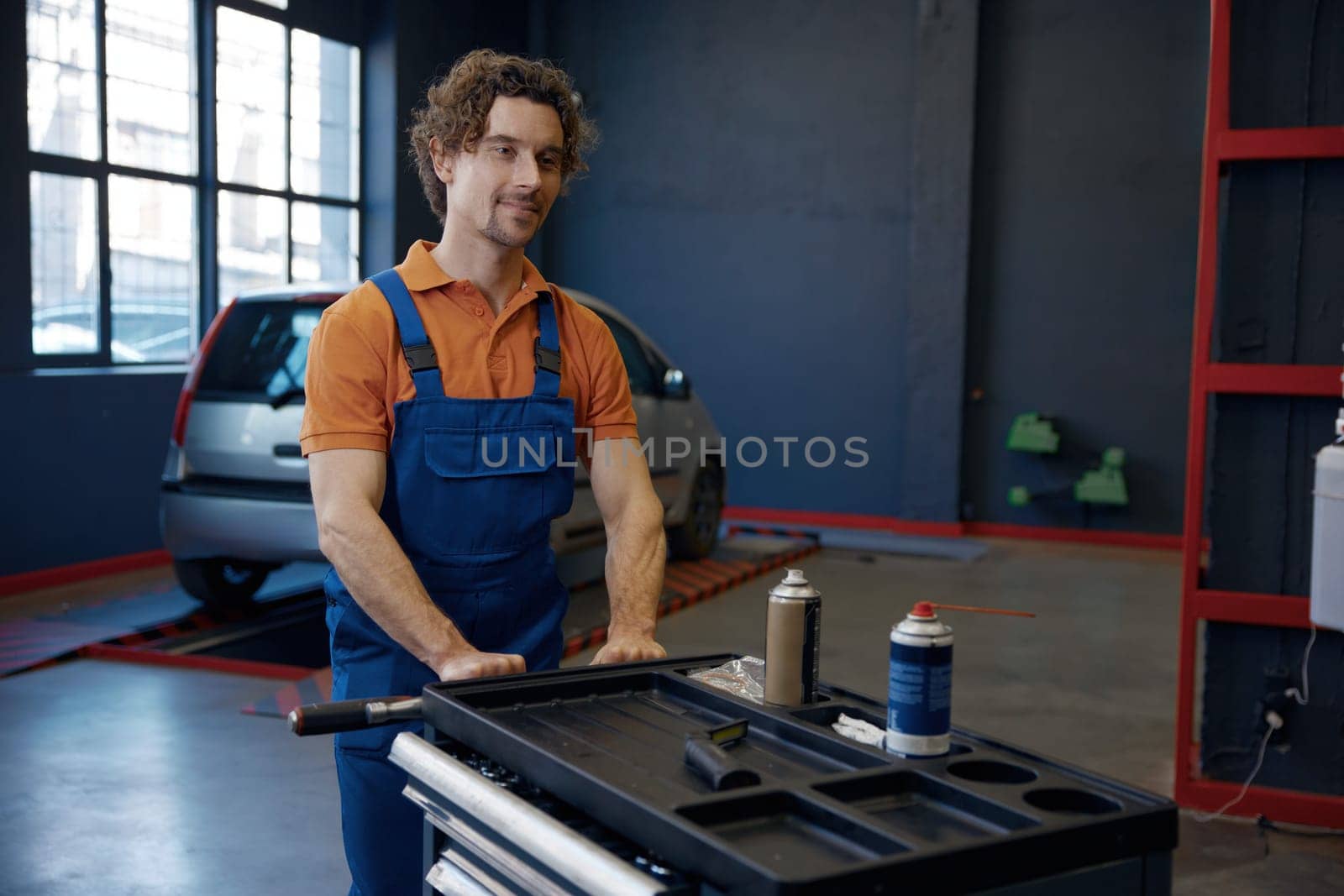 Young man mechanic working at car repair service workshop. Male technician pushing trolley cat with tools and supplies for auto maintenance and tuning