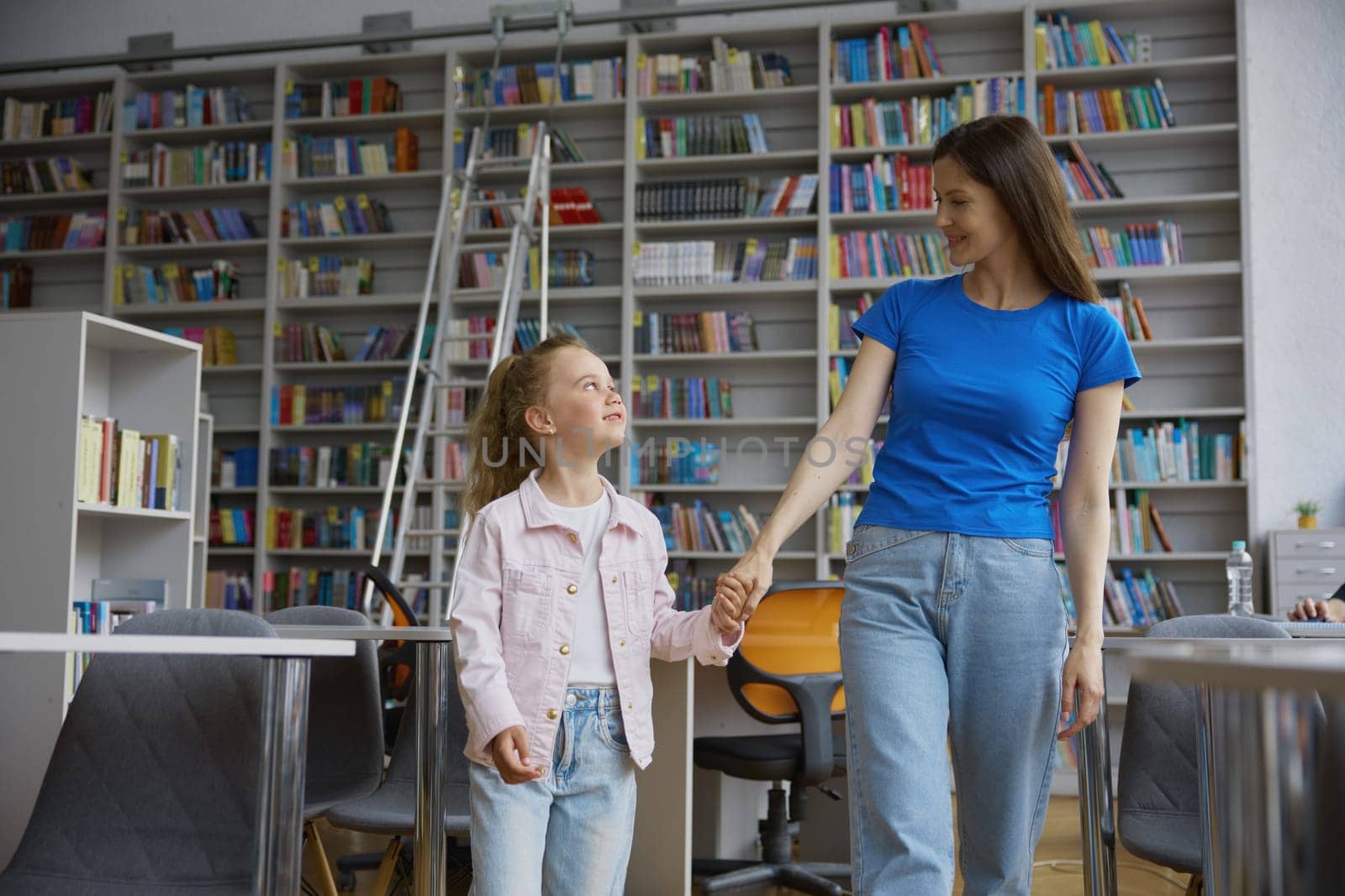Happy mother and daughter child standing over bookstore background. Mom and preschool kid holding hands looking at each other in anticipation of a well-spent time at library