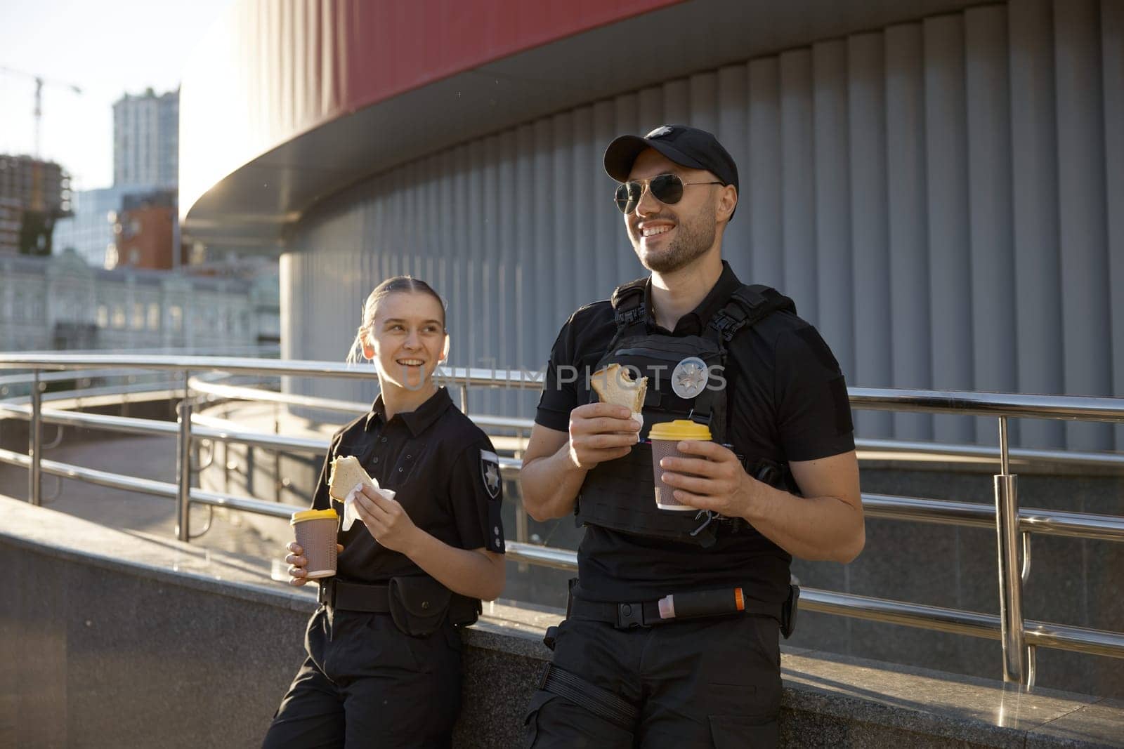 Male and female patrol police officers enjoying lunch break. Policeman and policewoman resting outdoor, drinking coffee, eating sandwich and having friendly conversation