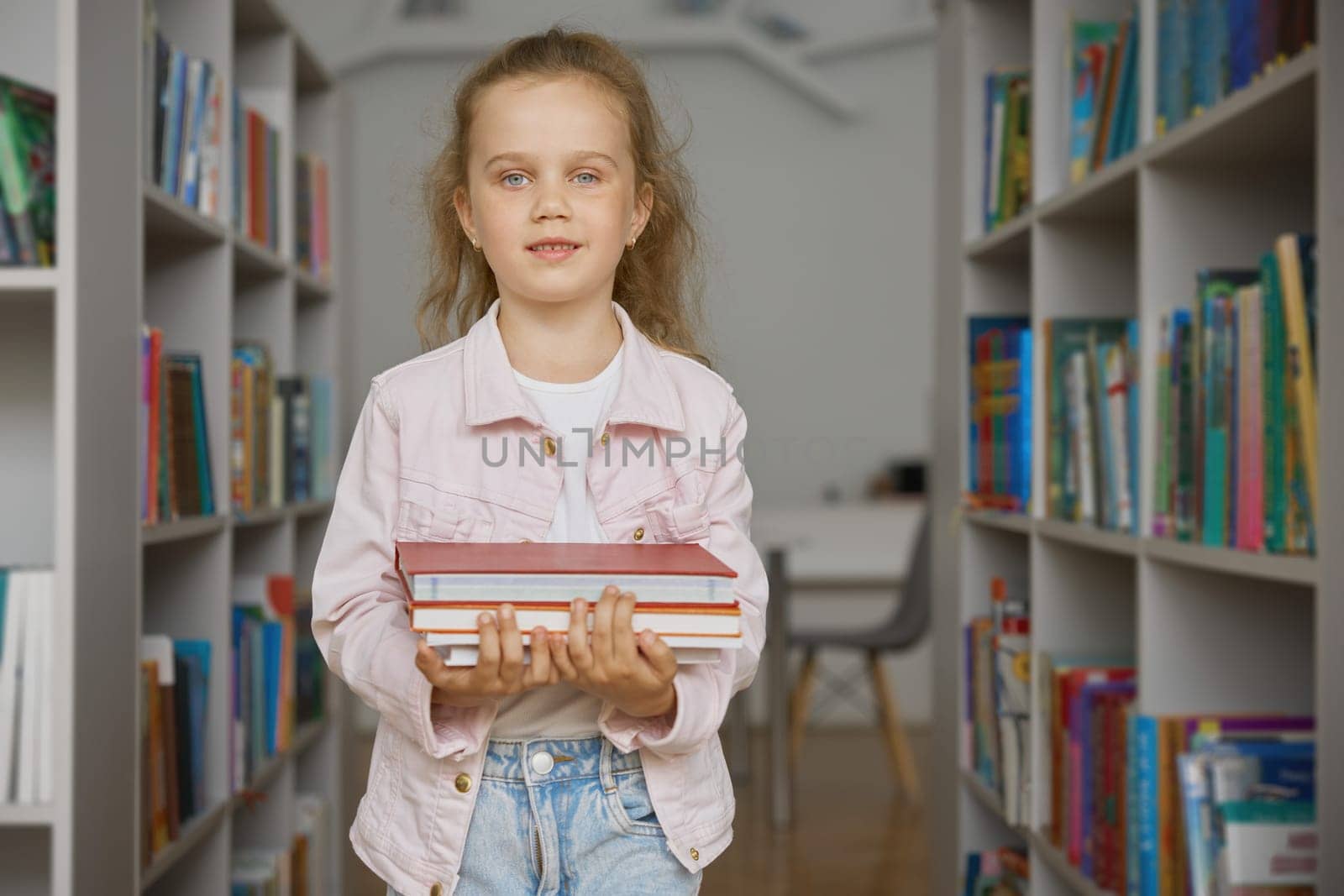 Portrait of little curious girl child carrying paper books stack in hands over bookstore interior background. Curiosity, knowledge and development of children