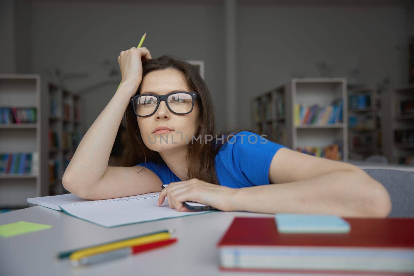 Bored young woman wearing eyeglasses drawing with pencils during homework preparation in library