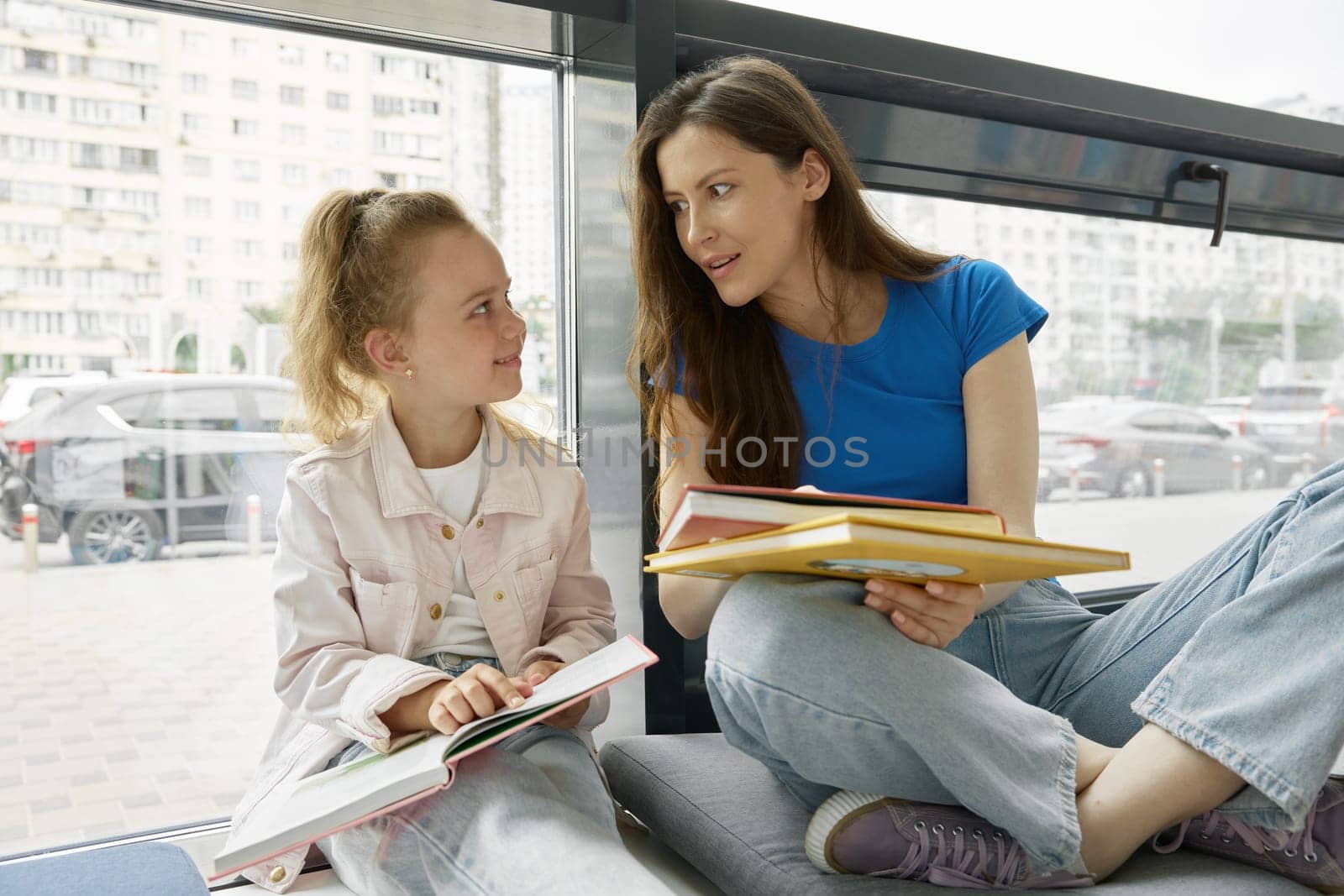 Mother and daughter reading books sitting on windowsill in city library. Family hobby to relax or be happy concept