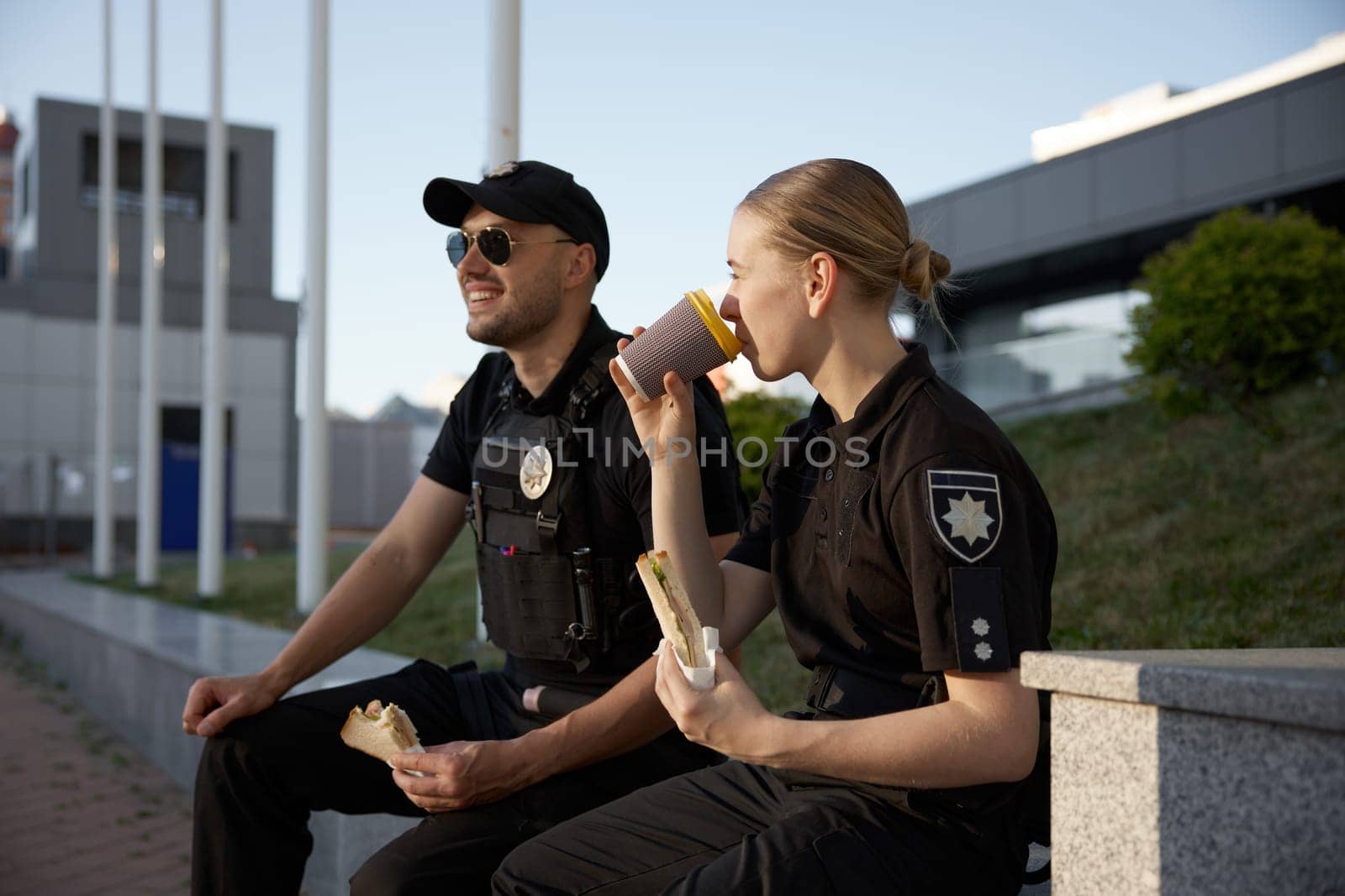 Patrol police officers enjoying lunch break. Policeman and policewoman resting outdoor, drinking coffee, eating sandwich and having friendly conversation