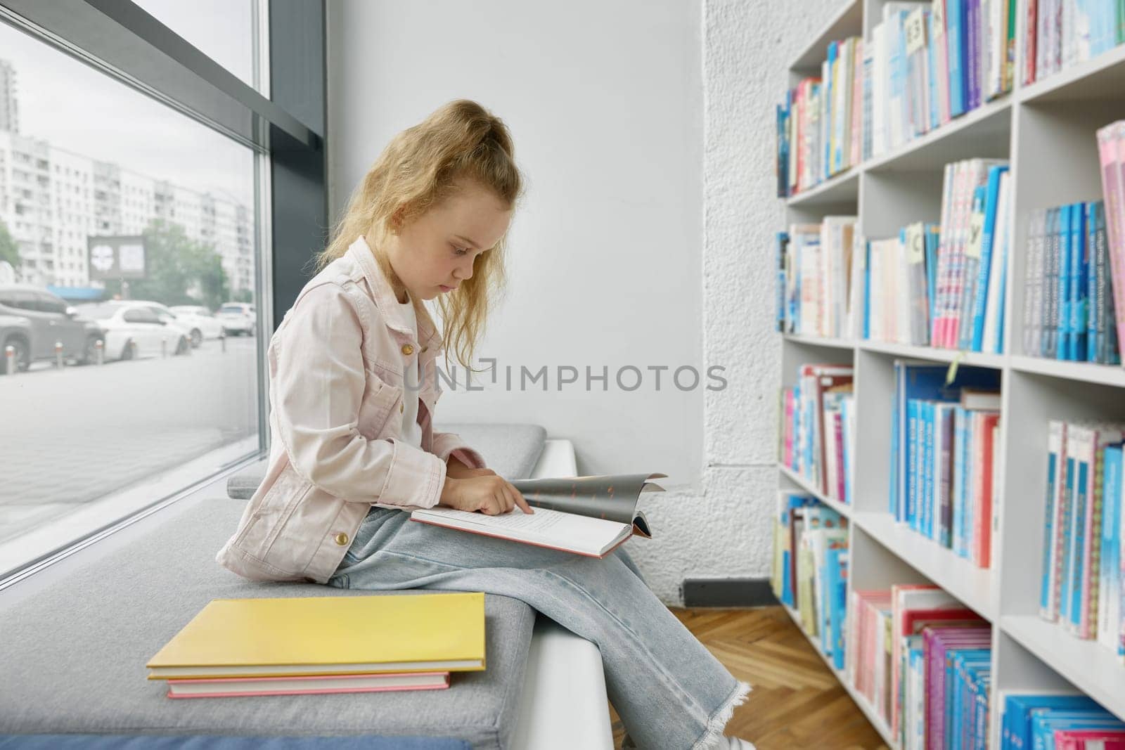 Pretty little girl child reading paper book while spending time in public library for children. Preschool female kid sitting on bench front of bookcase with shelves full of literature