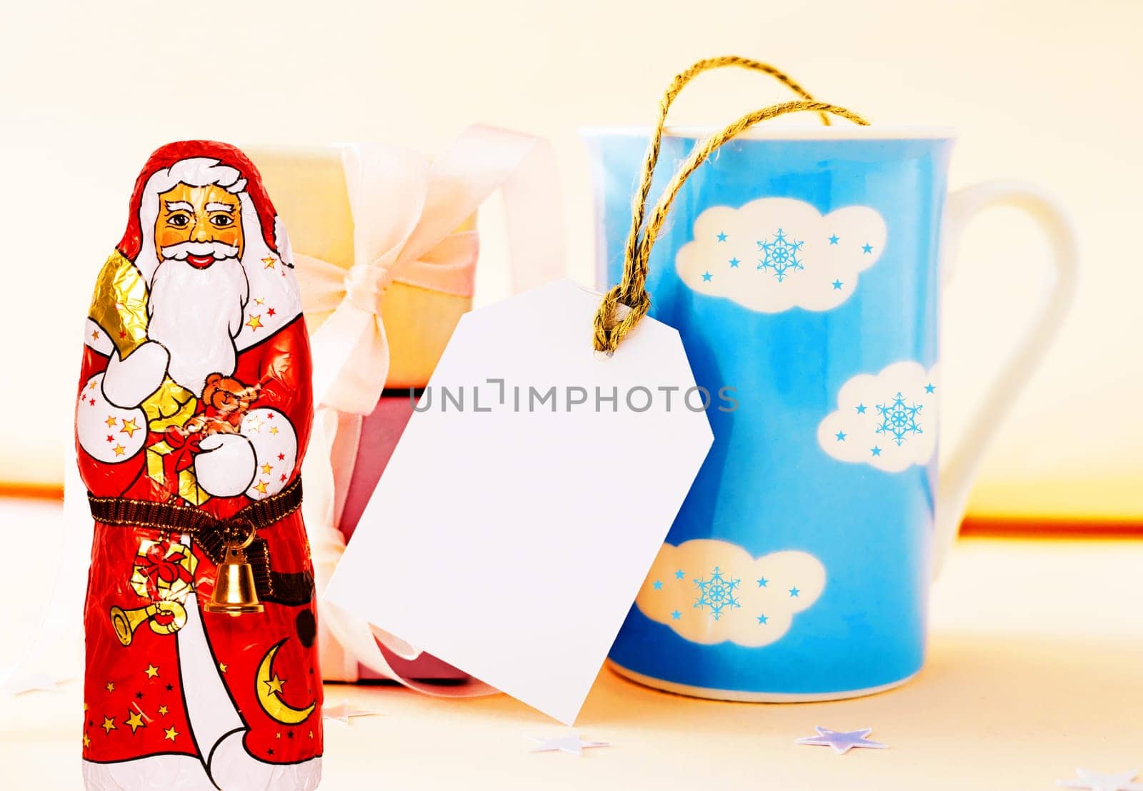 Christmas greeting card. Large blue white clouds winter mug with empty copy space tag, a cheerful wrapped chocolate Santa Claus and giftboxes on table over bright background.