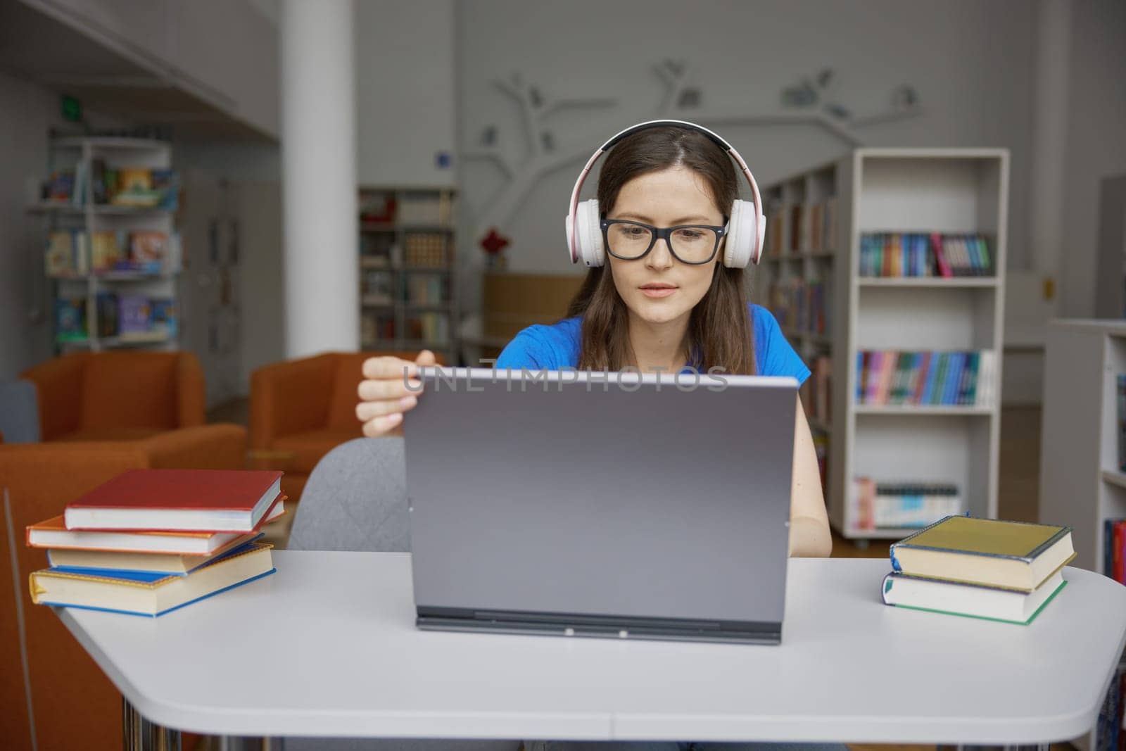 Front view portrait of smiling positive young woman studying with laptop in library. Teenage student wearing eyeglasses and headphones sitting at table desk looking at camera