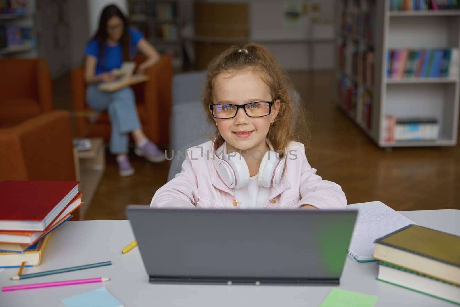 Cute happy girl child studying online via internet connection using laptop computer in public library while mother reading on blurred background. Portrait of smart little student