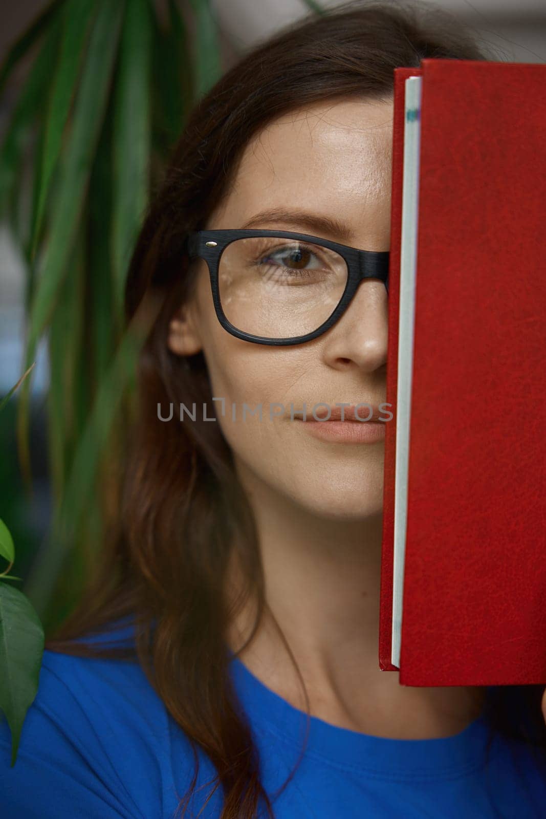 Closeup portrait of teenage girl student covering half part of face with book. Young pretty woman wearing eyeglasses looking at camera with smile