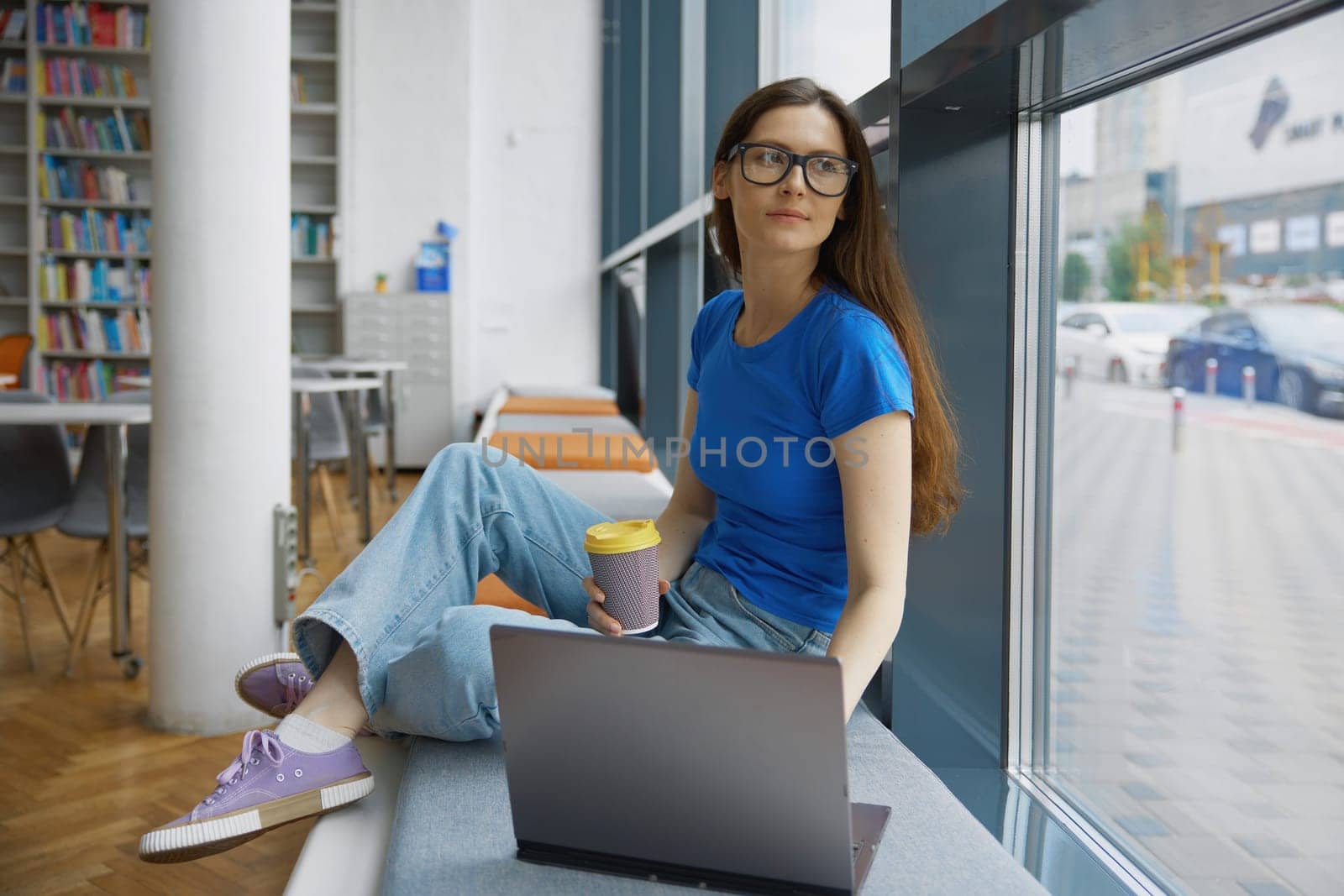 Young woman drinking coffee using laptop for studying in library. Pretty teenage student sitting on windowsill looking at computer screen enjoying aroma beverage browsing in internet at bookstore