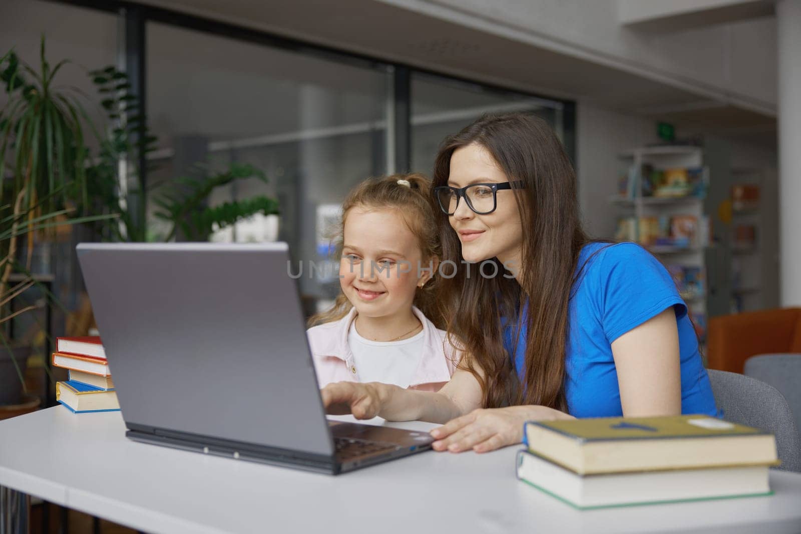 Young woman and girl kid using laptop for studying in library. Tutor and little preschool child reading paper book and online sources on internet