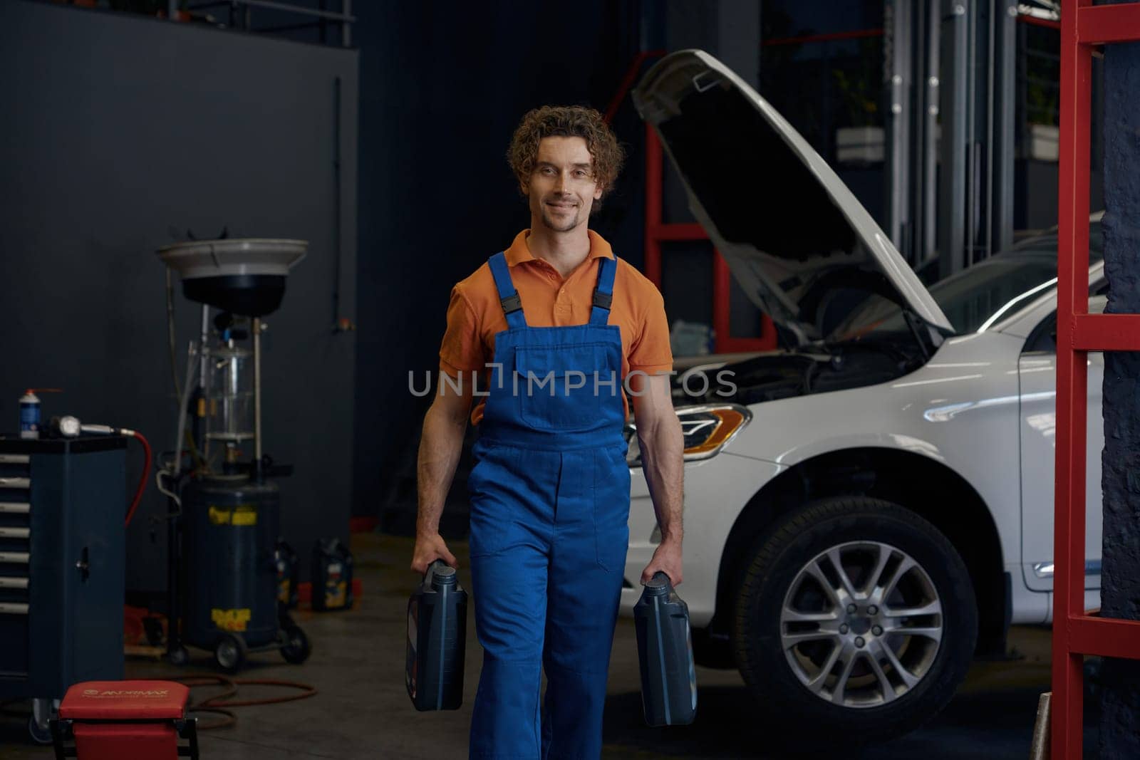 Man auto mechanic wearing overalls carrying canisters with car motor oil walking to camera. Work at automotive service shop