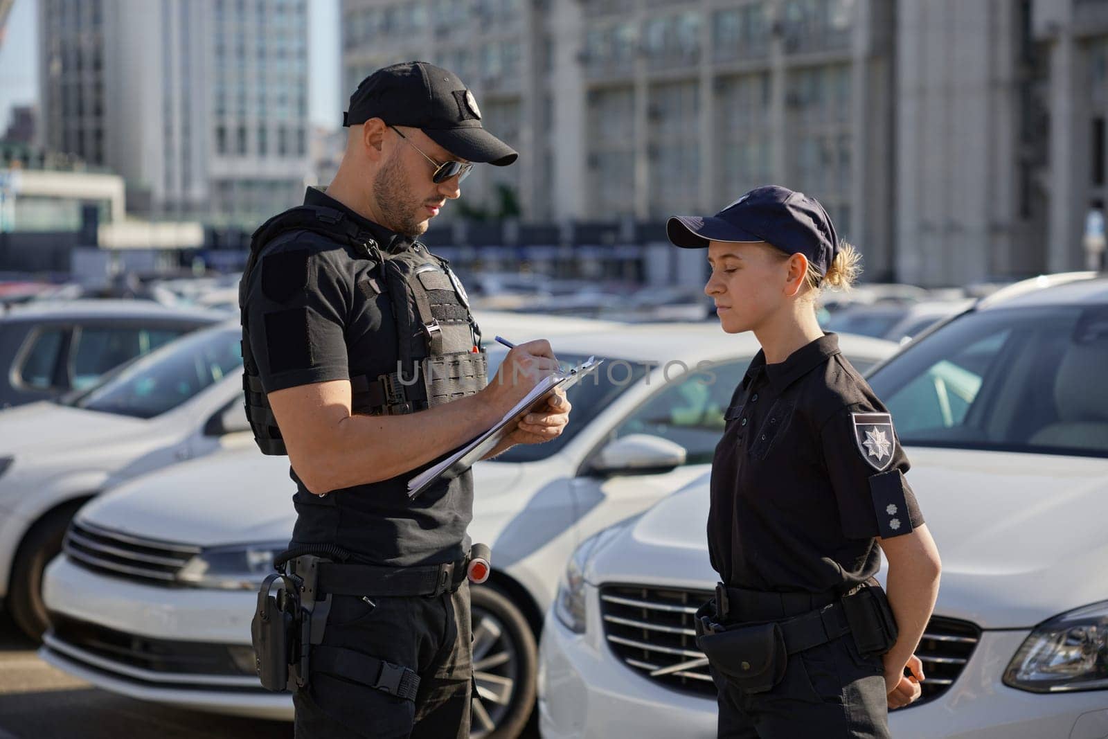 Woman and man patrol police team working on city street with parked cars. Policeman writing in clipboard, teaching young policewoman