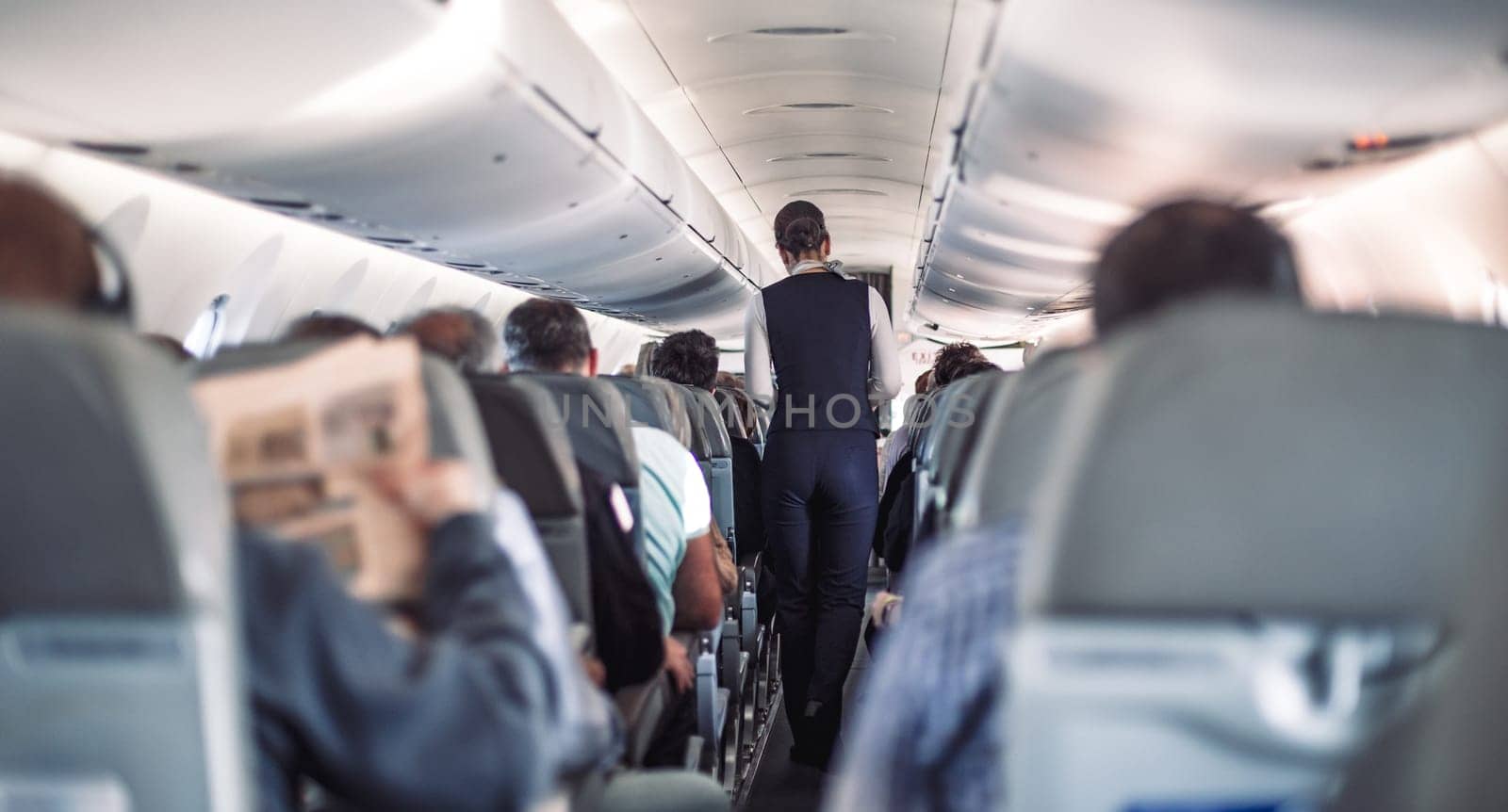 Interior of airplane with passengers on seats and stewardess in uniform walking the aisle, serving people. Commercial economy flight service concept