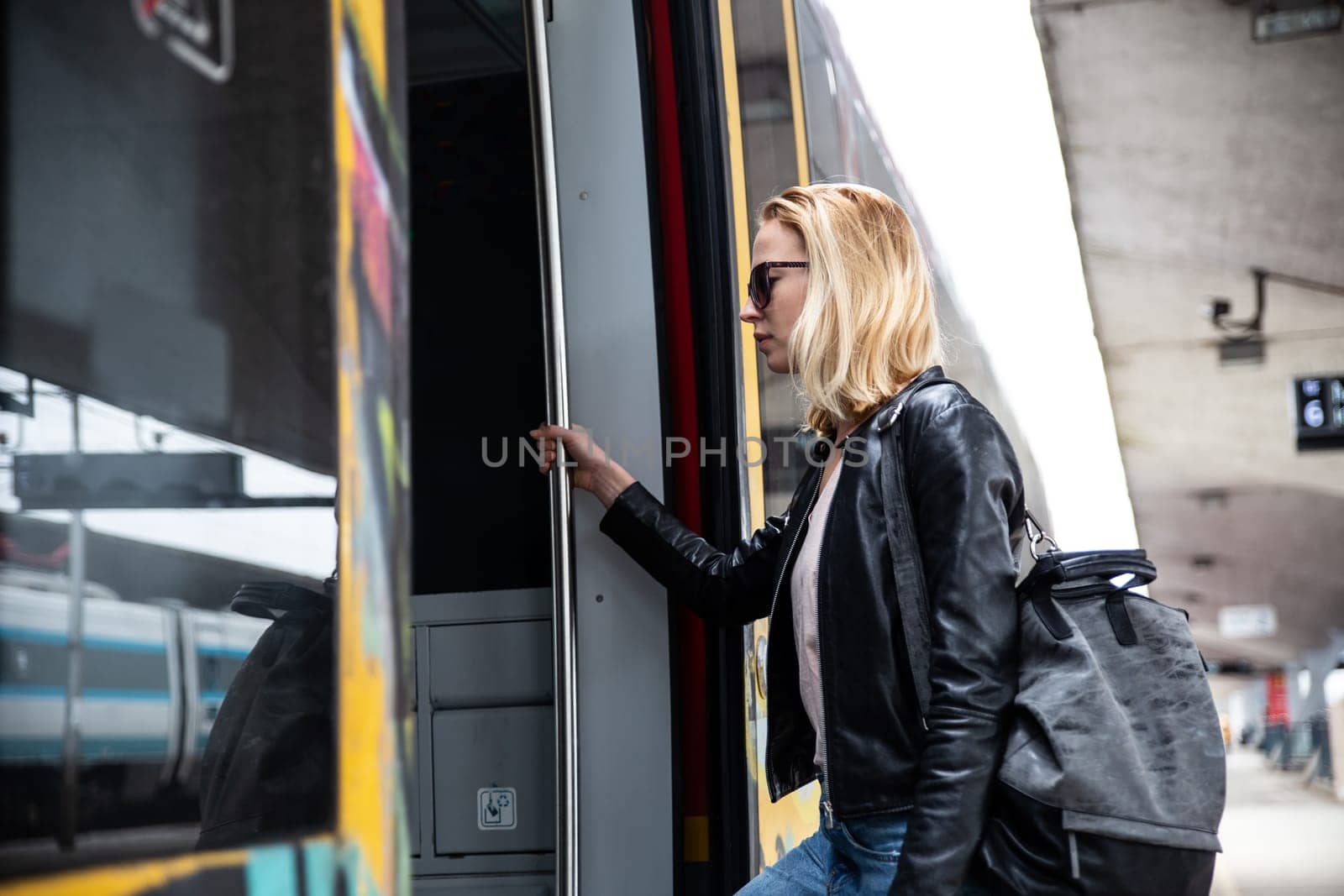Young blond woman in jeans, shirt and leather jacket wearing bag and sunglass, presses door button of modern speed train to board on train station platform. Travel and transportation.