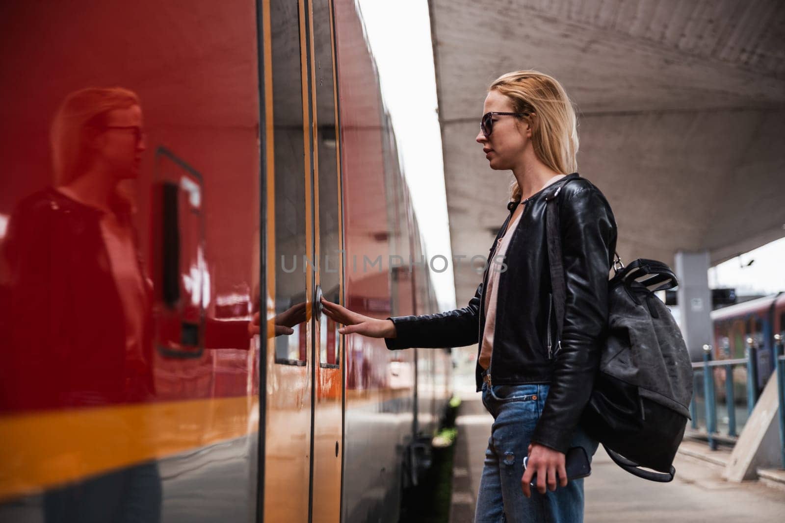 Young blond woman in jeans, shirt and leather jacket wearing bag and sunglass, presses door button of modern speed train to board on train station platform. Travel and transportation.