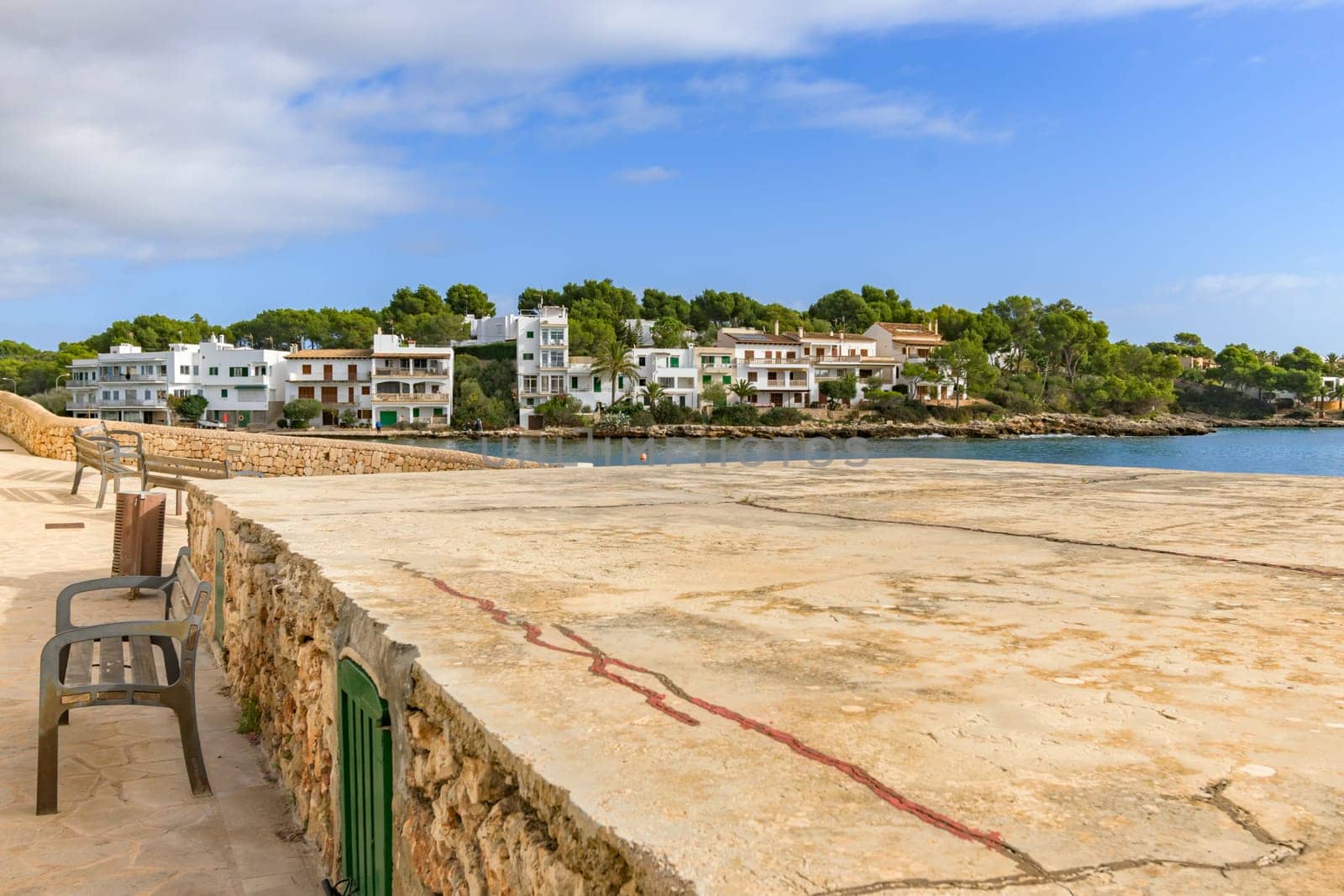 Coastal town with modern buildings and a stone promenade under a partly cloudy sky, porto petro, mallorca balearic islands, spain