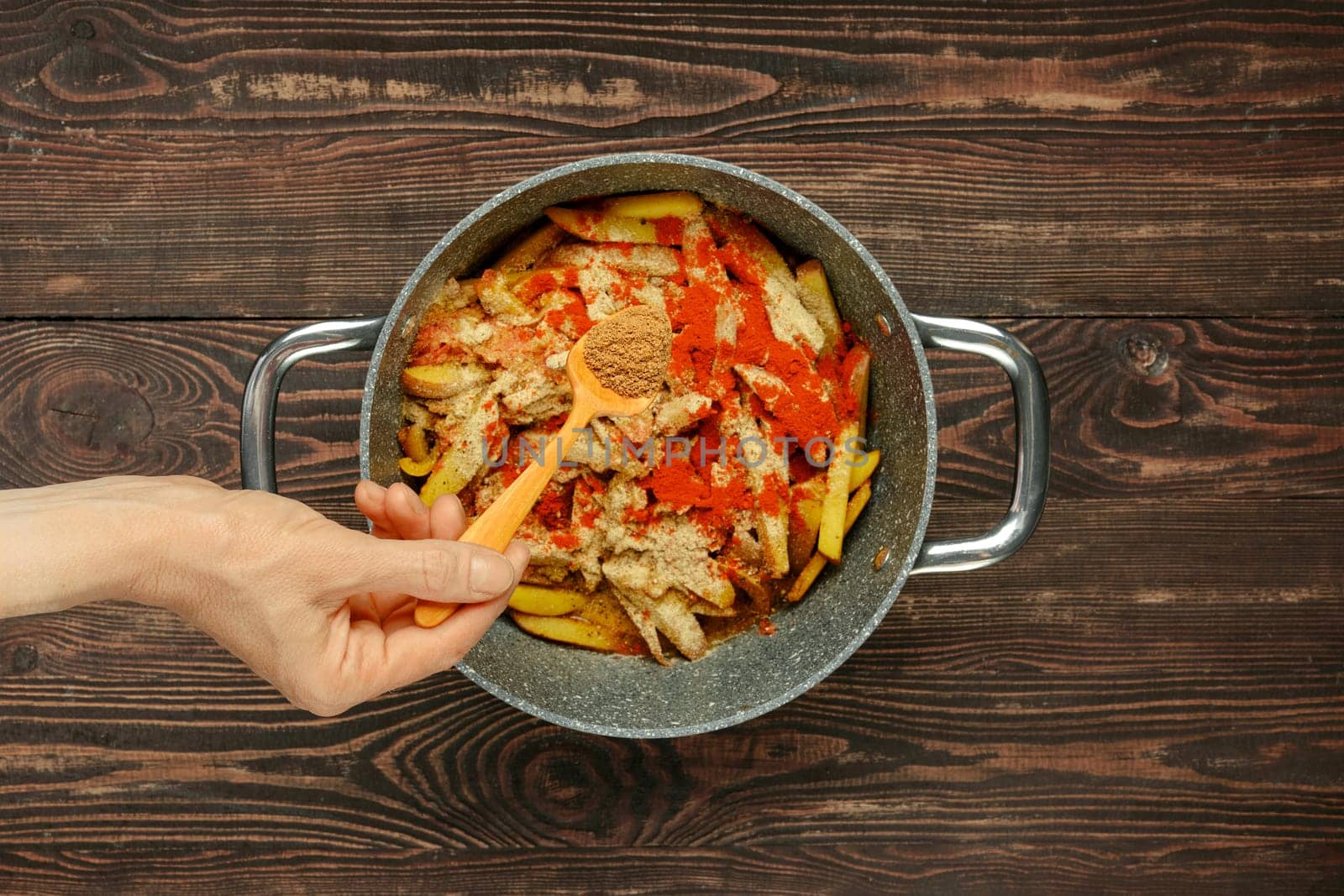 A hand adds coriander to a pot filled with sliced potatoes and bell peppers, preparing a flavorful dish on a rustic wooden table. The vibrant ingredients suggest a hearty, home-cooked meal.