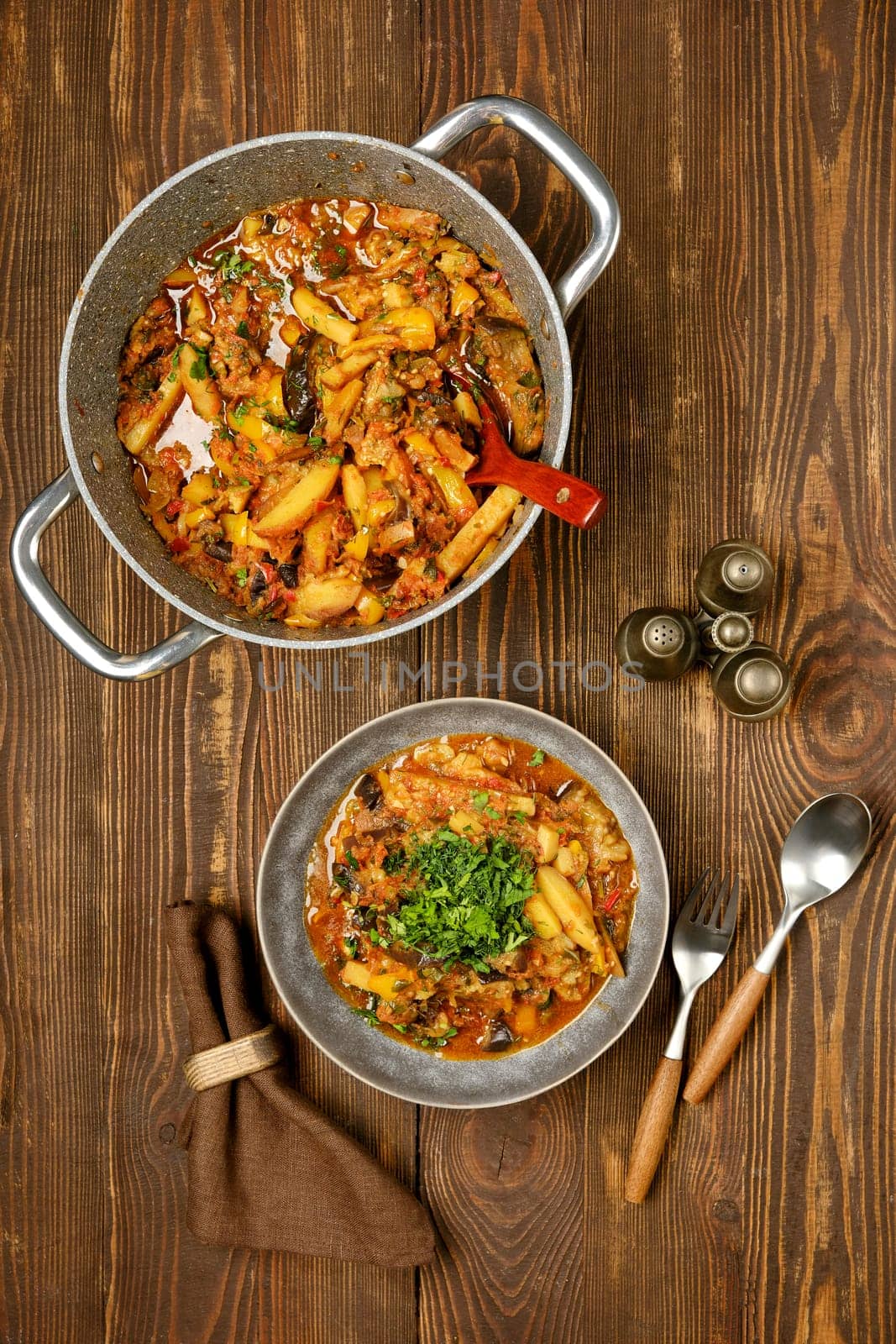 Top view of a warm vegetable stew, rich with various ingredients in a bowl beside a pot. Fresh herbs garnish the dish, creating an inviting meal on a rustic wooden surface.