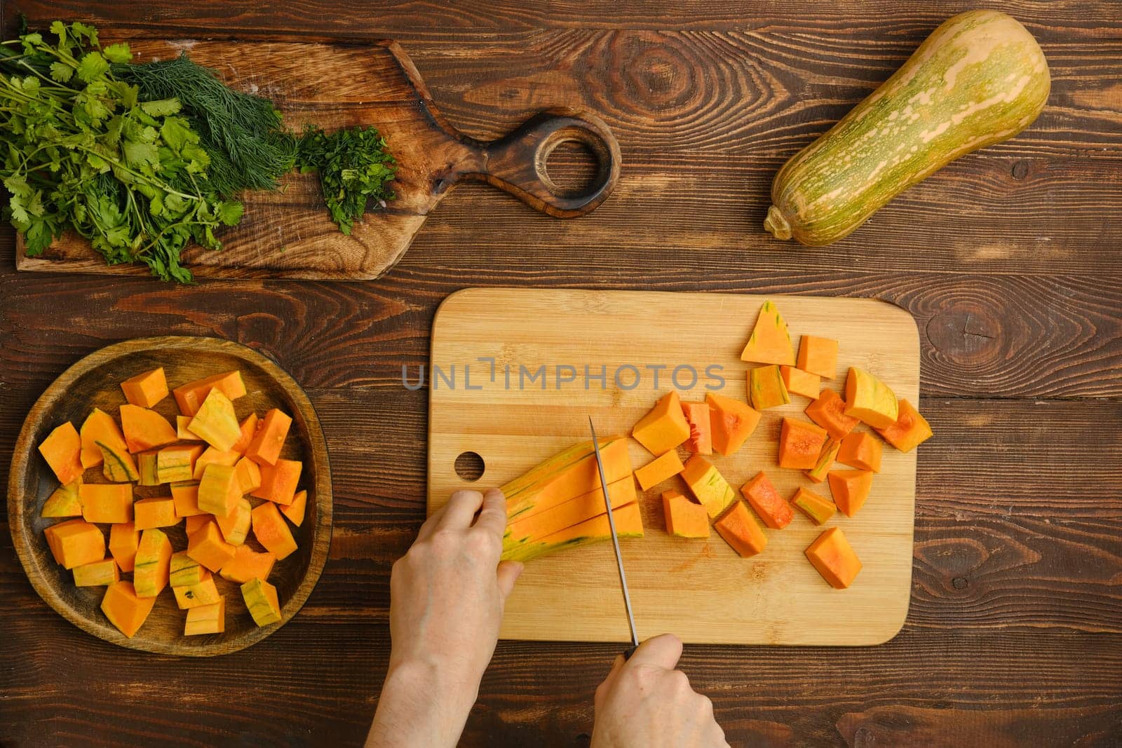 A person is skillfully chopping vibrant orange squash into cubes on a wooden cutting board. Fresh herbs are laid out beside them, creating a warm kitchen atmosphere filled with culinary inspiration.