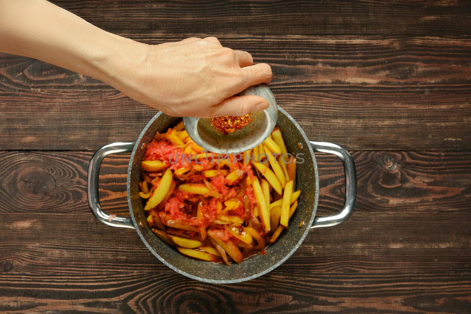 A hand sprinkles chili pepper flakes over a pan filled with fresh tomatoes and potatoes in a warm kitchen. The wooden countertop adds a rustic touch, enhancing the inviting atmosphere of home cooking.