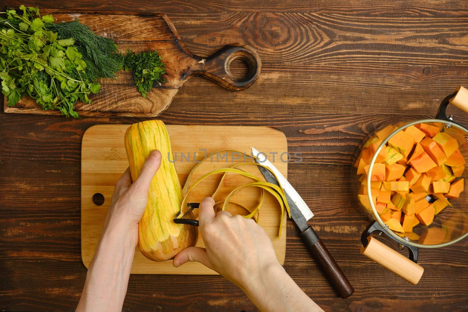 A person is peeling fresh squash with a vegetable peeler, surrounded by freshly chopped herbs and diced squash in a pot, creating a warm and inviting atmosphere in a home kitchen.