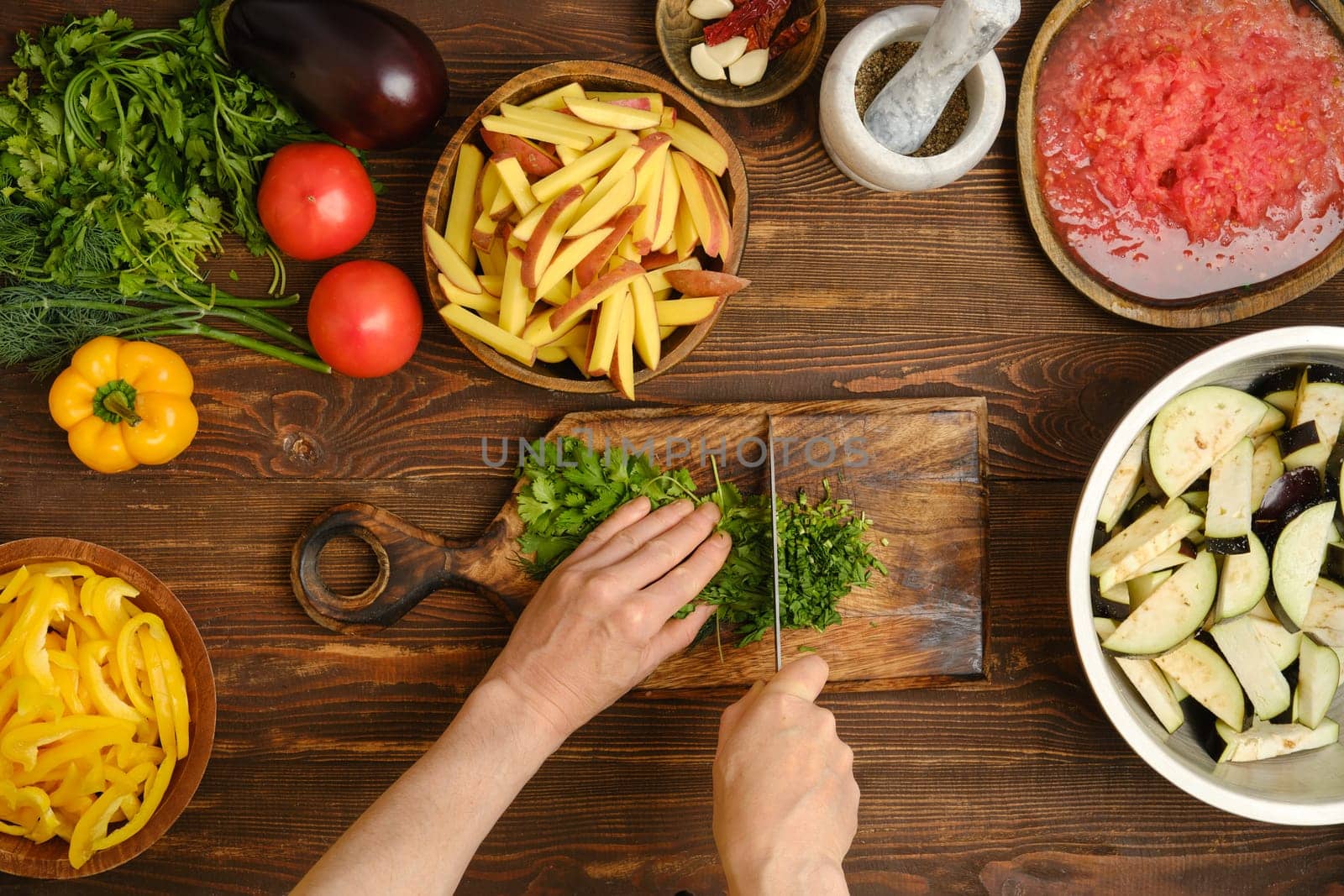 A person chops fresh cilantro on a wooden cutting board while surrounded by various colorful vegetables and green herbs in a warm kitchen. The preparations emphasize a focus on healthy cooking.