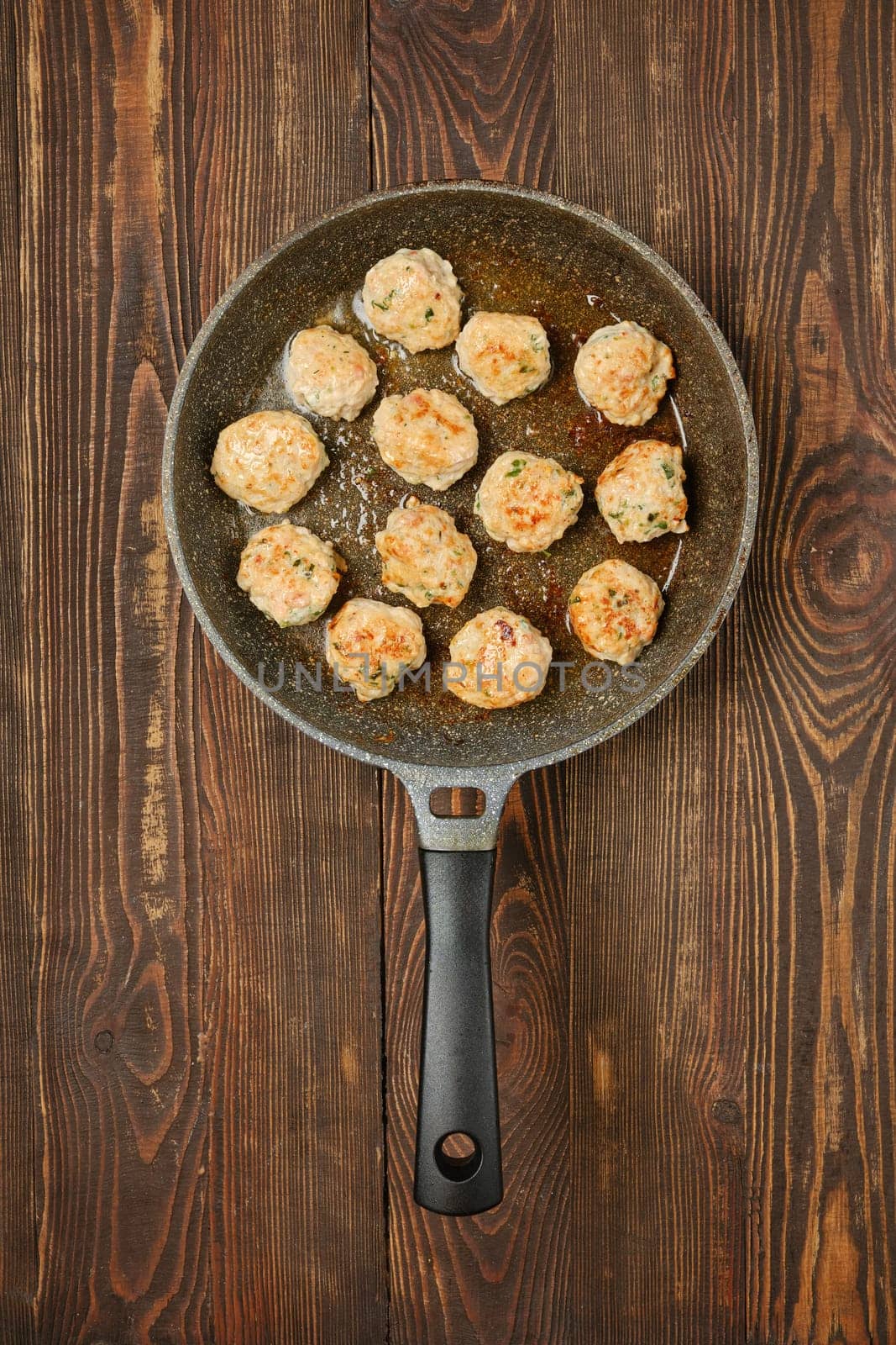 Meatballs are sizzling in a skillet on a rustic wooden table, showcasing a delicious cooking process. The dish highlights various spices and herbs, contributing to a flavorful meal preparation.