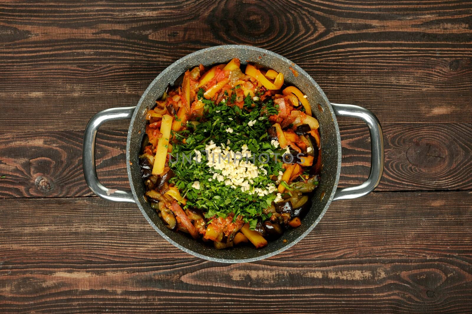 A colorful stir-fry of bell peppers, eggplant, and potatoes, topped with fresh herbs and garlic, sits in a sturdy pan on a rustic wooden table.