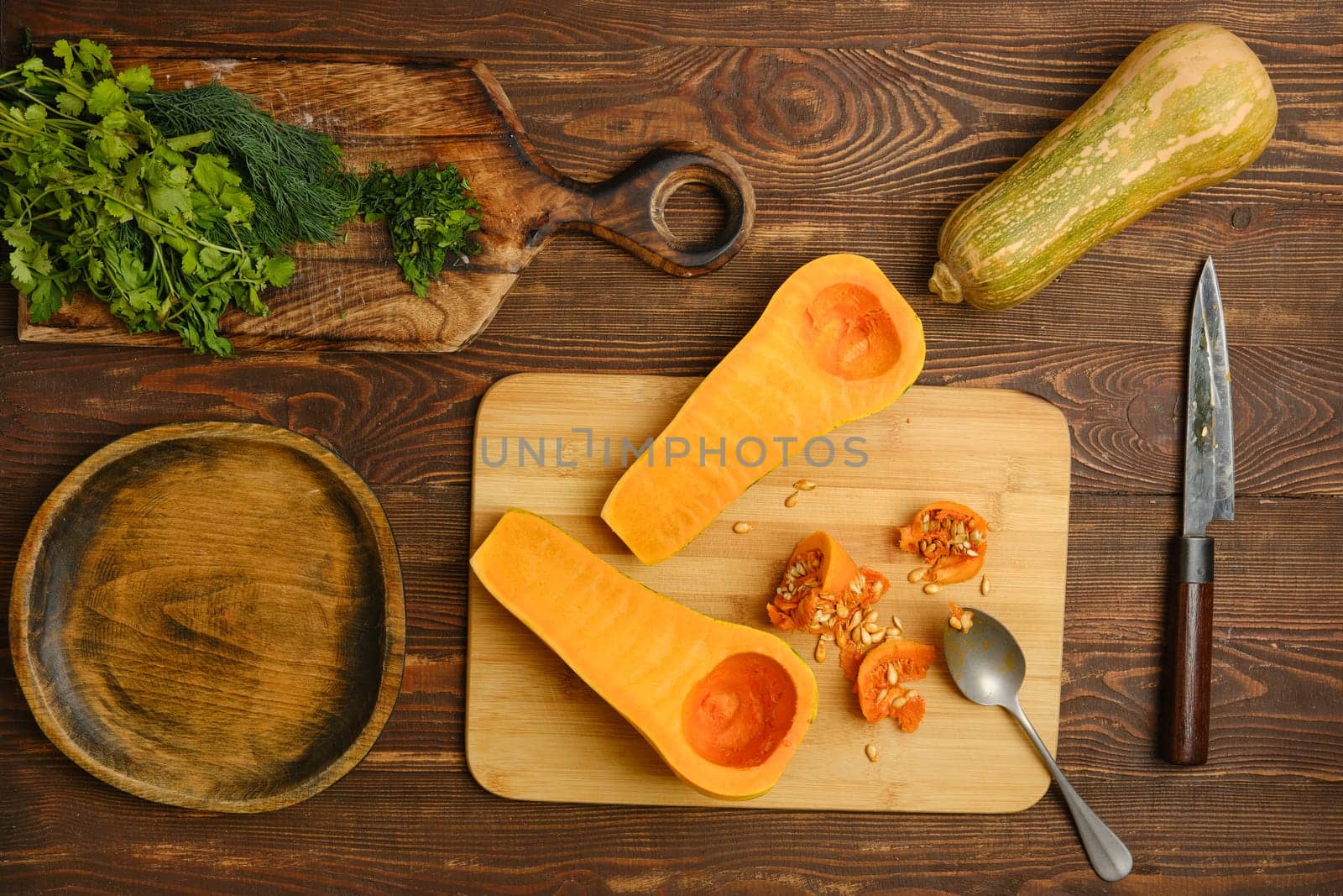 A halved butternut squash on a wooden cutting board alongside fresh herbs. Nearby, a knife and spoon are placed, while a whole squash and a wooden bowl complete the kitchen setting.
