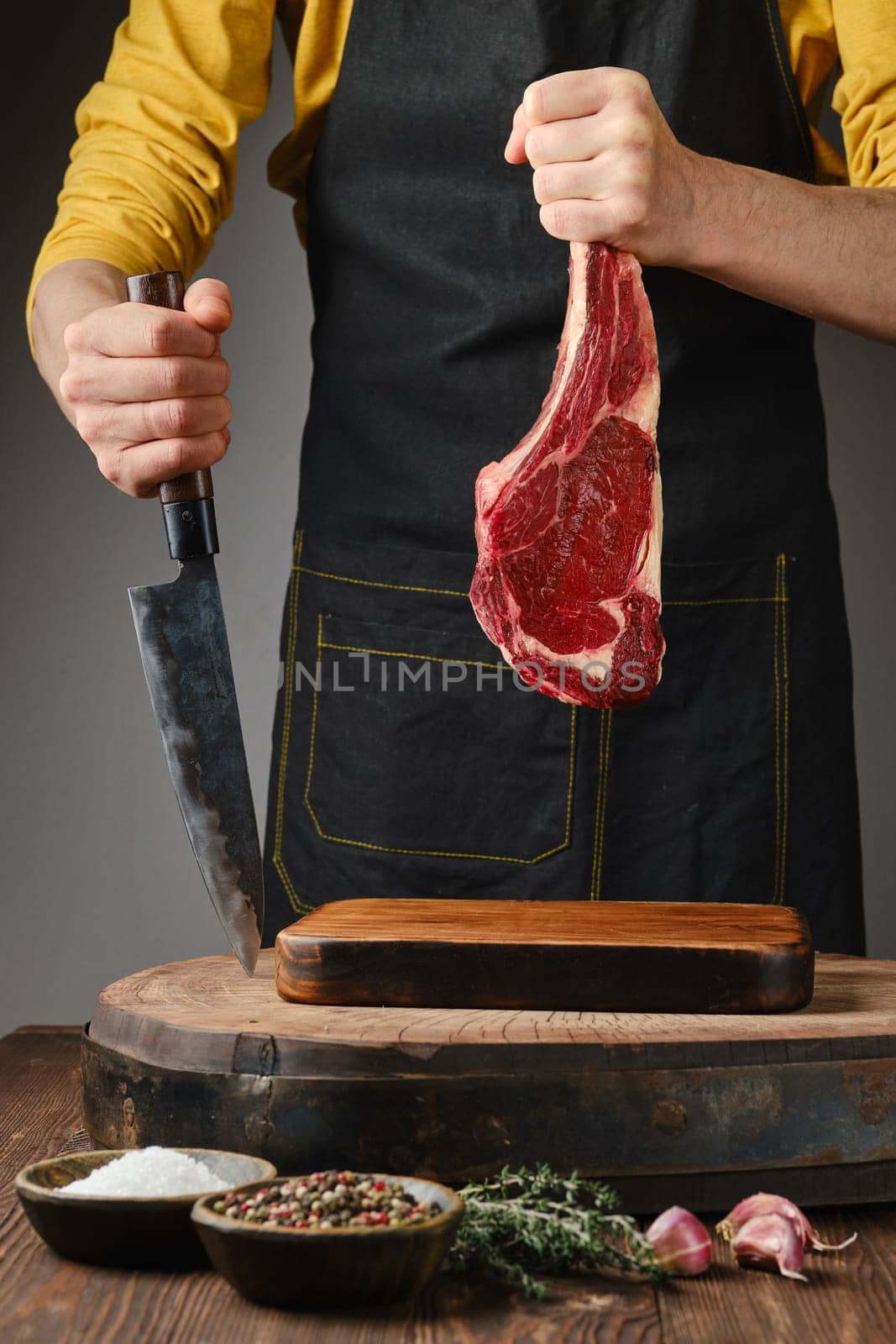 A person in a yellow shirt and apron holds a large cut of raw meat while preparing to slice it on a wooden board. Fresh herbs and spices are arranged nearby, enhancing the culinary experience.