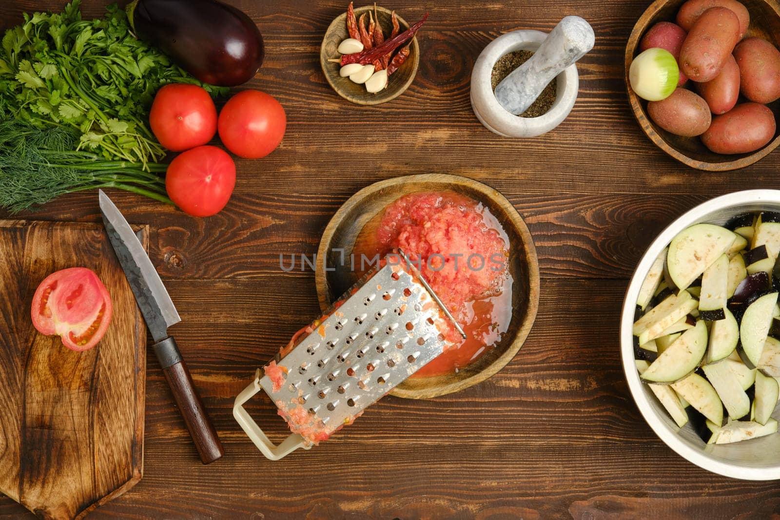 Grated fresh tomatoes and metal grater on a wooden bowl surrounded with eggplant, green herbs and potatoes, are arranged on a wooden table.