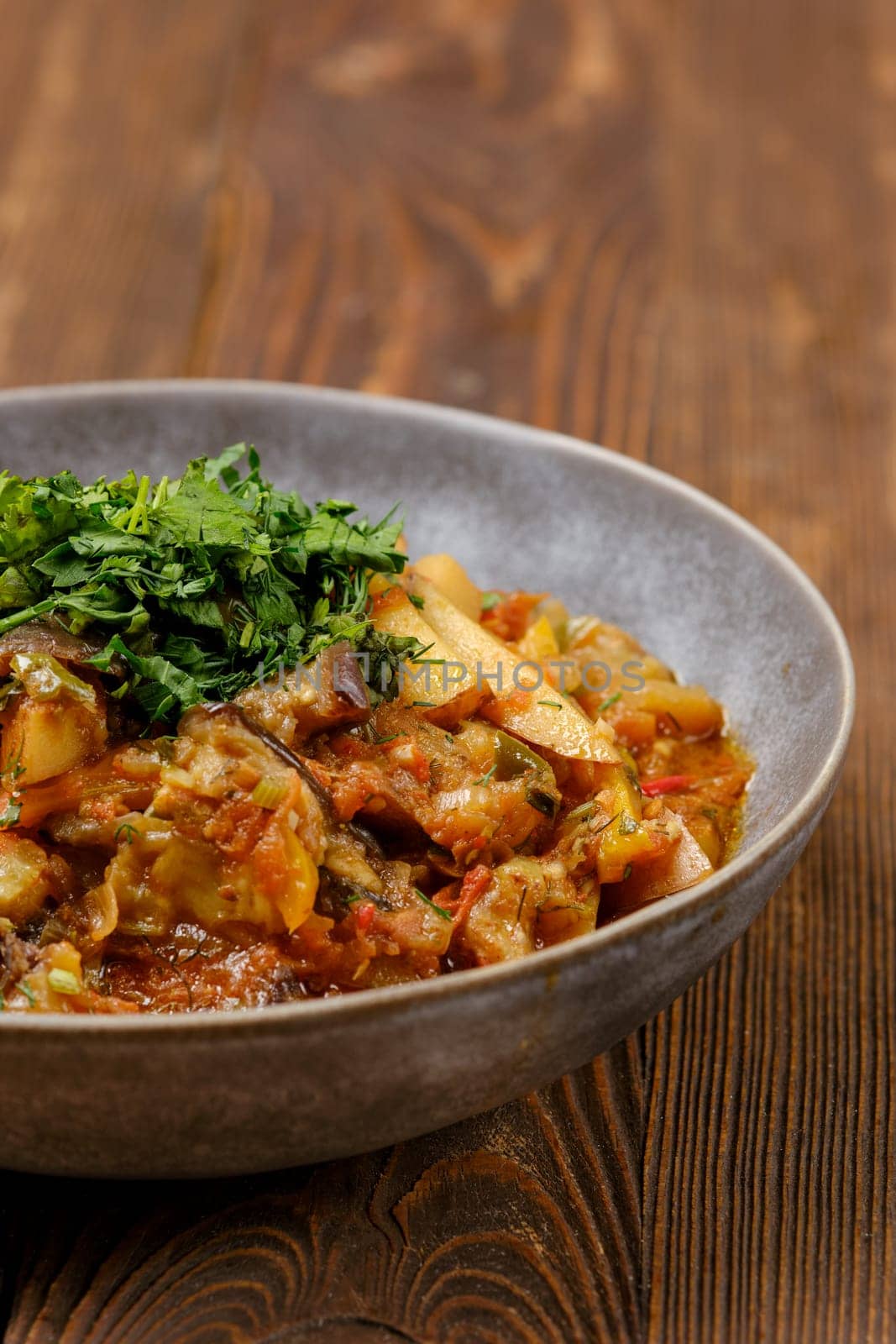 Close-up view of a vegetable stew filled with assorted vegetables, topped with fresh green herbs, served in a rustic bowl on a wooden table. The warm colors create an inviting and appetizing display.