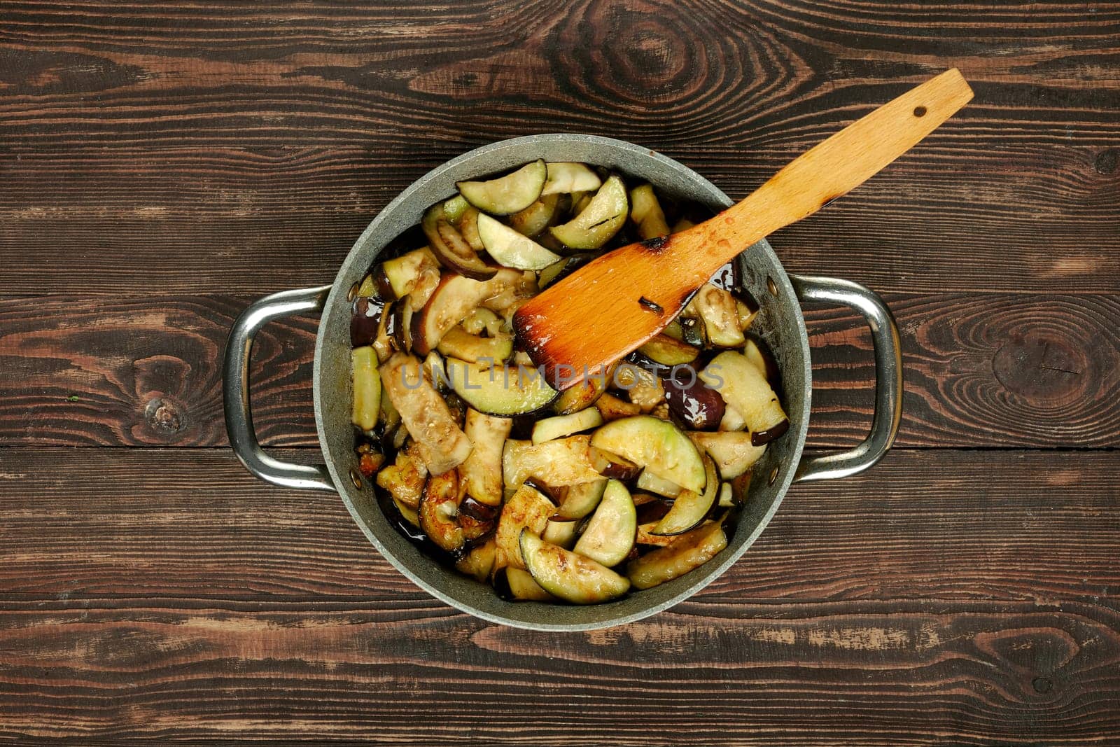 A skillet filled with sauteed zucchini and eggplant sits on a wooden countertop, with a wooden spatula resting inside, showcasing a warm and inviting cooking atmosphere.