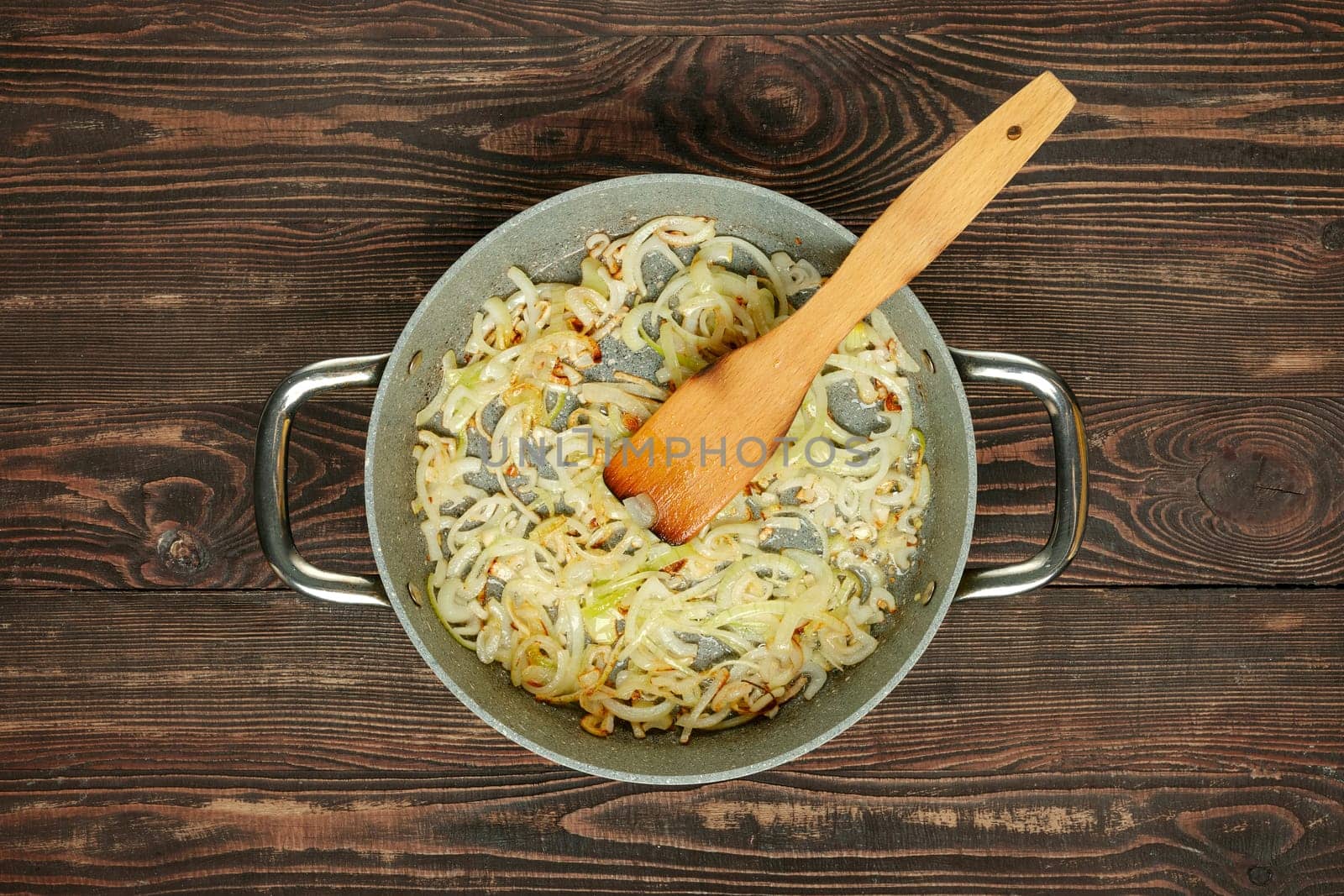 Chopped onions and garlic are sauteed in a pan with a wooden spatula, creating a flavorful base for a meal on a rustic wooden table. The scene captures the essence of home cooking.