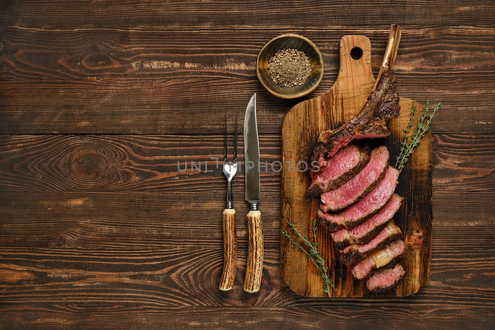 Succulent slices of tomahawk steak are arranged neatly on a rustic wooden cutting board, accompanied by fresh herbs and utensils. The scene captures a delicious meal prepared with care.
