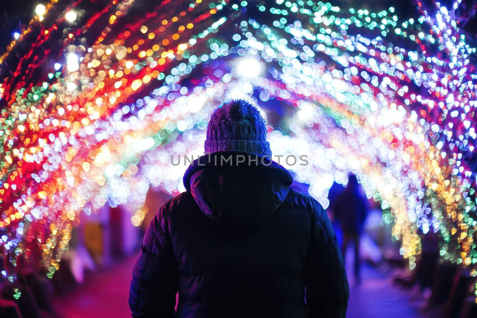 A person stands under a vibrant display of colorful lights enjoying the festive atmosphere of a winter festival in the evening.
