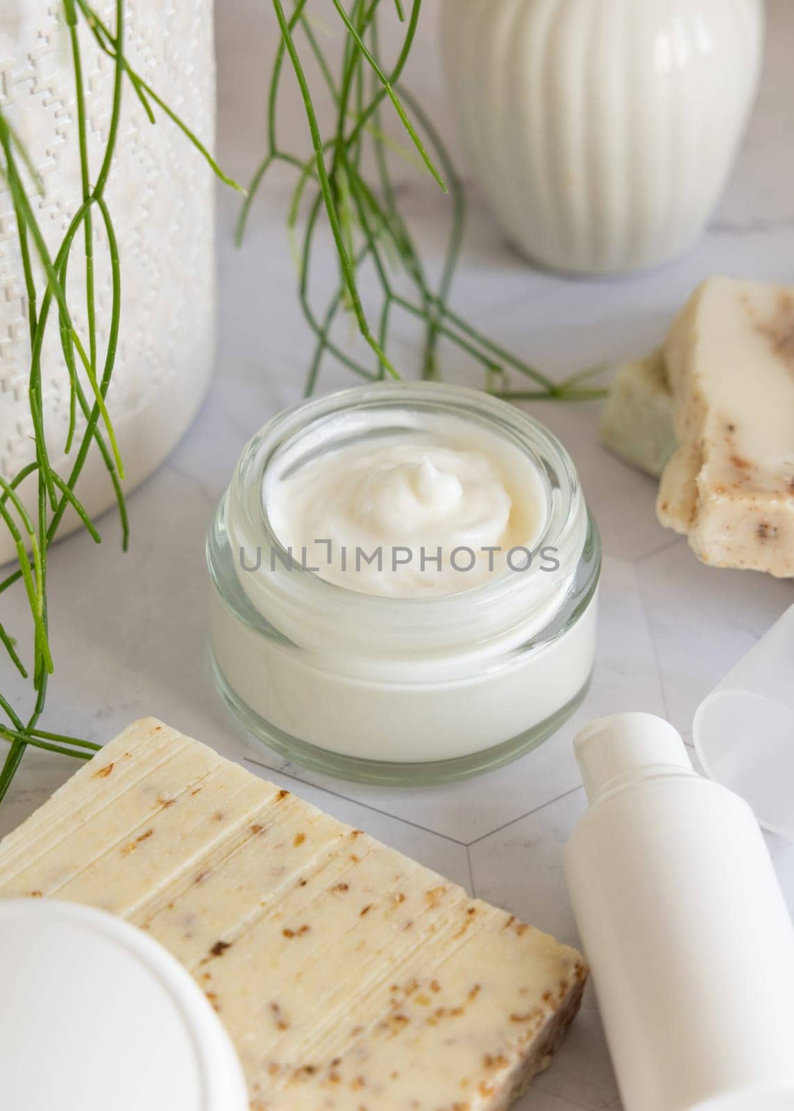 Opened cream jar with white blank lid on white marble against potted green plant and soap bars in bathroom, close up, packaging mockup. Cosmetic product for facecare, beauty routine