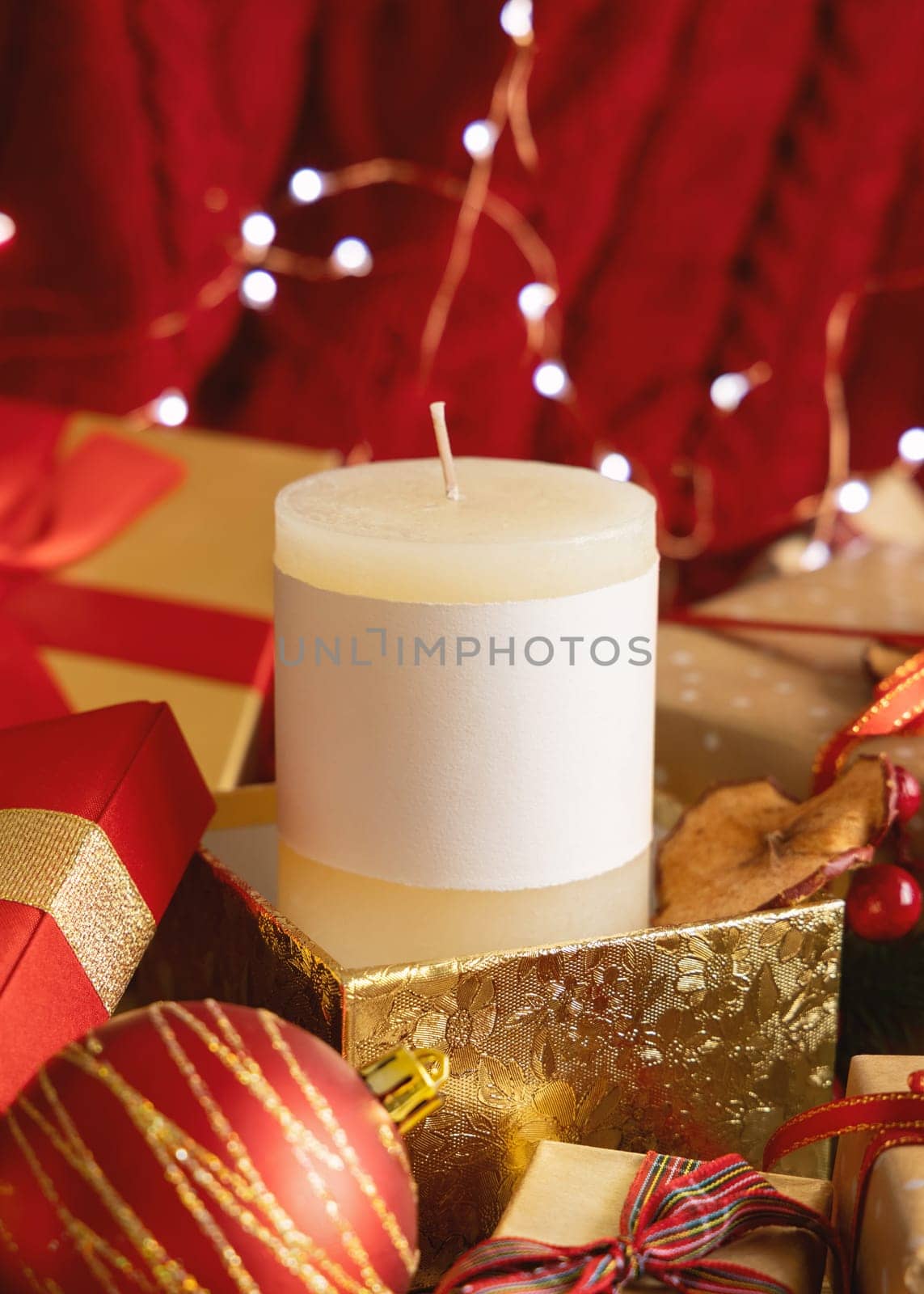 Pillar candle with blank label in golden Christmas gift box against red knitted sweater and lights, copy space. Warm winter family atmospheric mock up, Holiday time